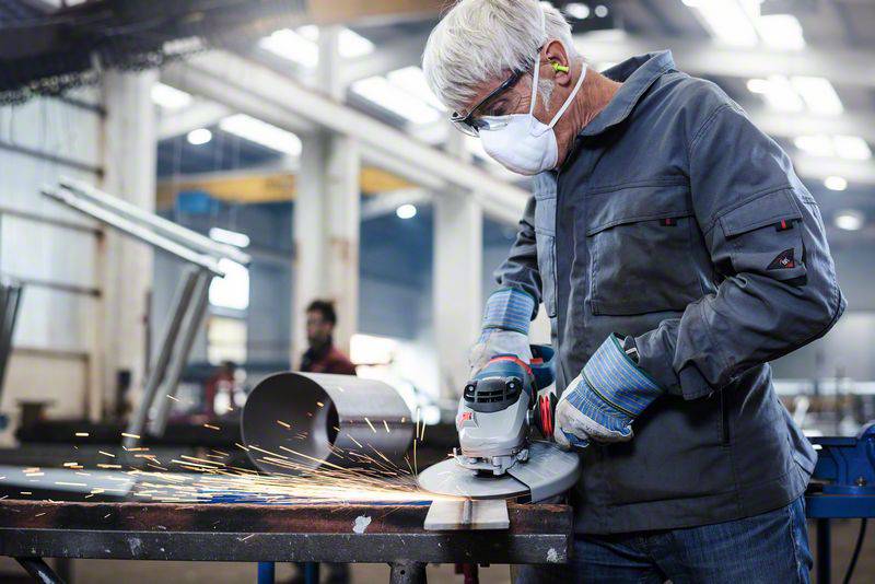 Ein Arbeiter mit Schutzbrille und Maske schleift Metall mit einem Winkelschleifer in einer Fabrik. Funken fliegen vom Werkstück.