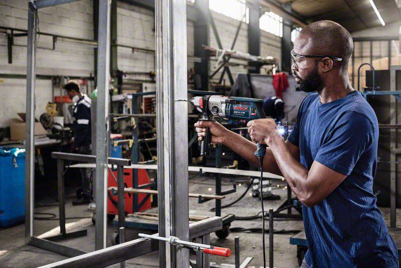 Ein Mann bohrt in einem Industriegebäude ein Metallgestell. Im Hintergrund arbeitet eine weitere Person an einem Tisch.