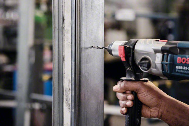Eine Person bohrt mit einer Bohrmaschine ein Loch in ein Metallstück. Gespanntes Gesicht, Fokus auf die Arbeit in einer Werkstatt.
