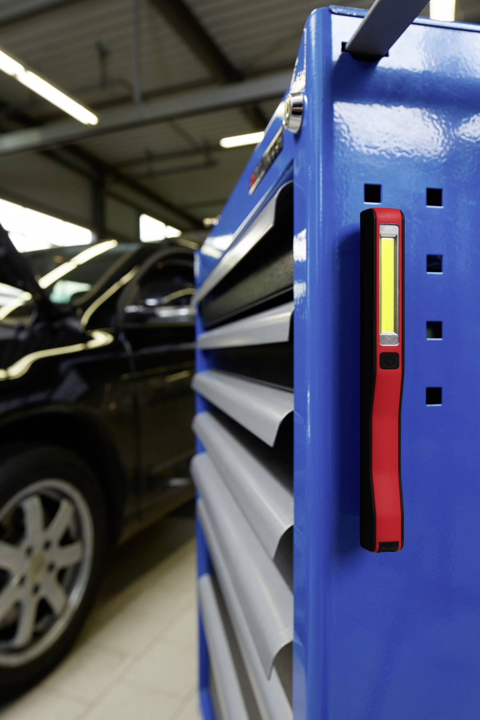 Blauer Werkstattwagen mit Werkzeug, rotes Magnetlicht befestigt. Im Hintergrund steht ein Auto mit geöffneter Motorhaube in einer Werkstatt.