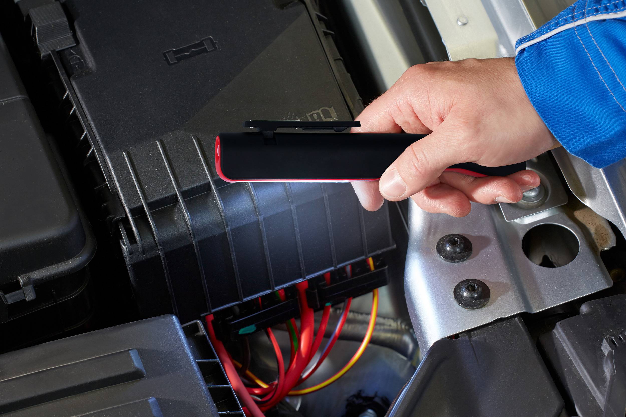 Eine Person hält eine tragbare Starthilfe über die Autobatterie eines Autos, bereit zur Anwendung. Kabel und Motorkomponenten sind sichtbar.