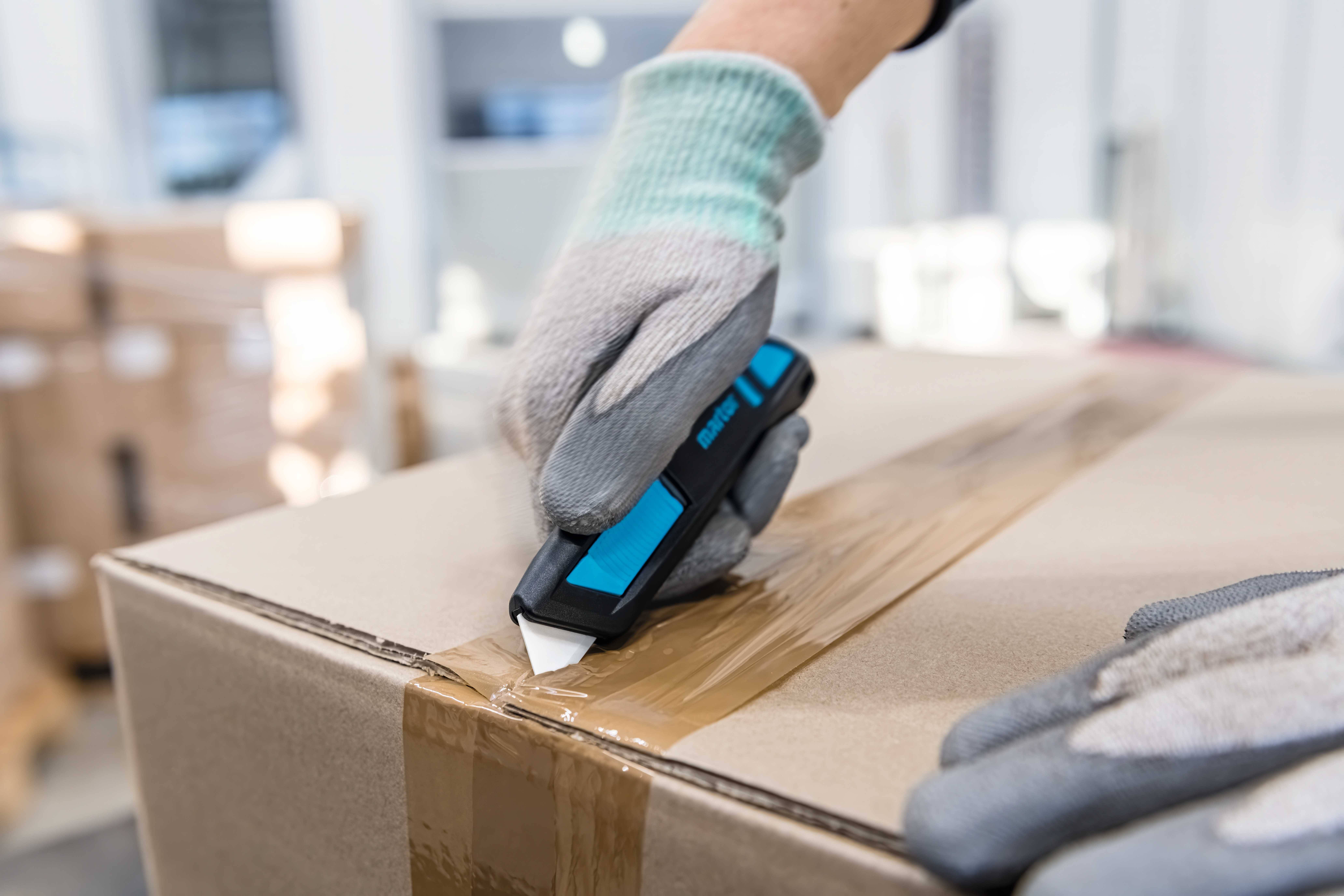 Person in Handschuhen öffnet mit einem Cuttermesser einen Karton. Der Karton ist mit Klebeband versiegelt und befindet sich in einem Lager.