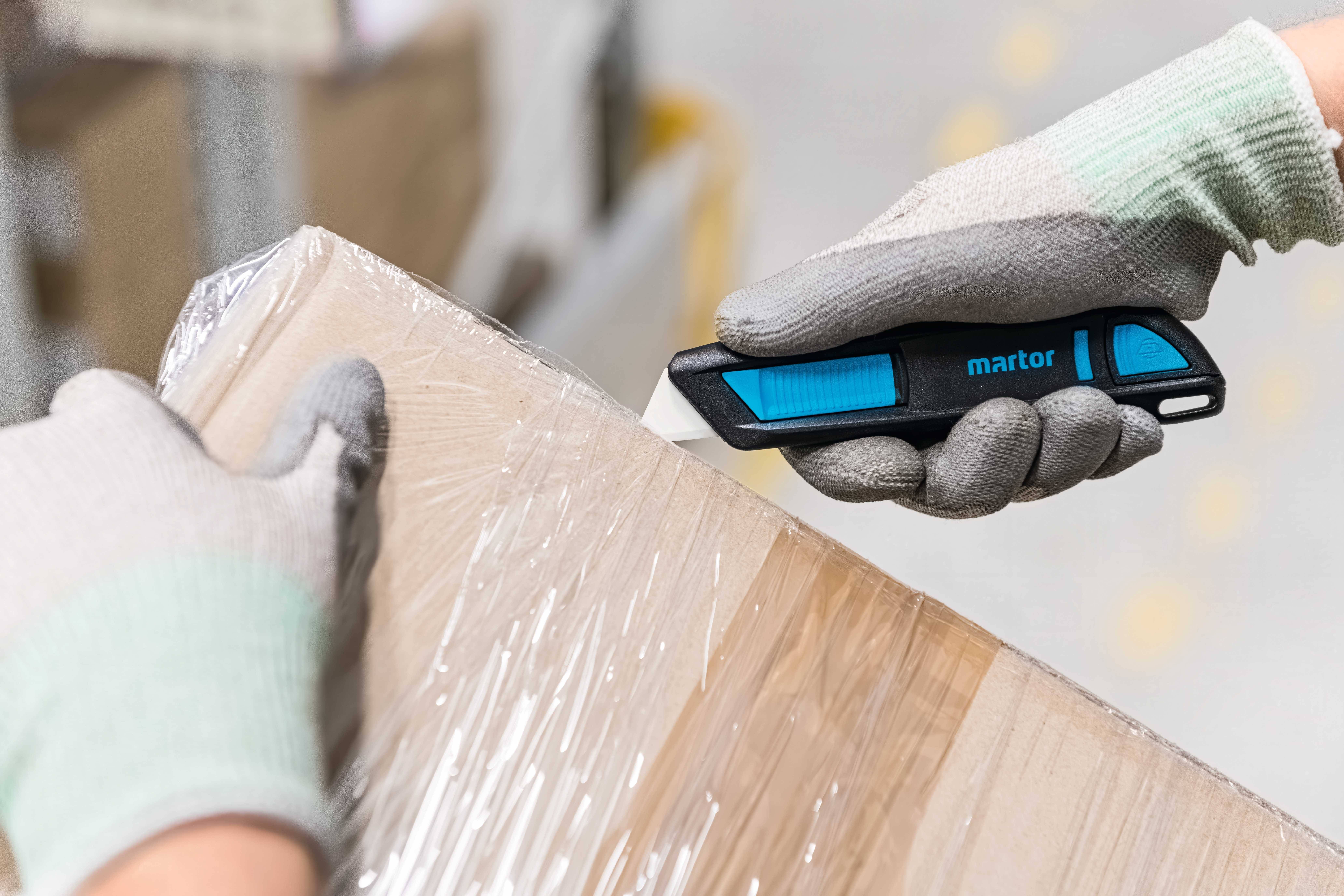 Zwei behandschuhte Hände schneiden mit einem blauen Sicherheitsmesser eine verpackte, mit Folie umwickelte Box auf.