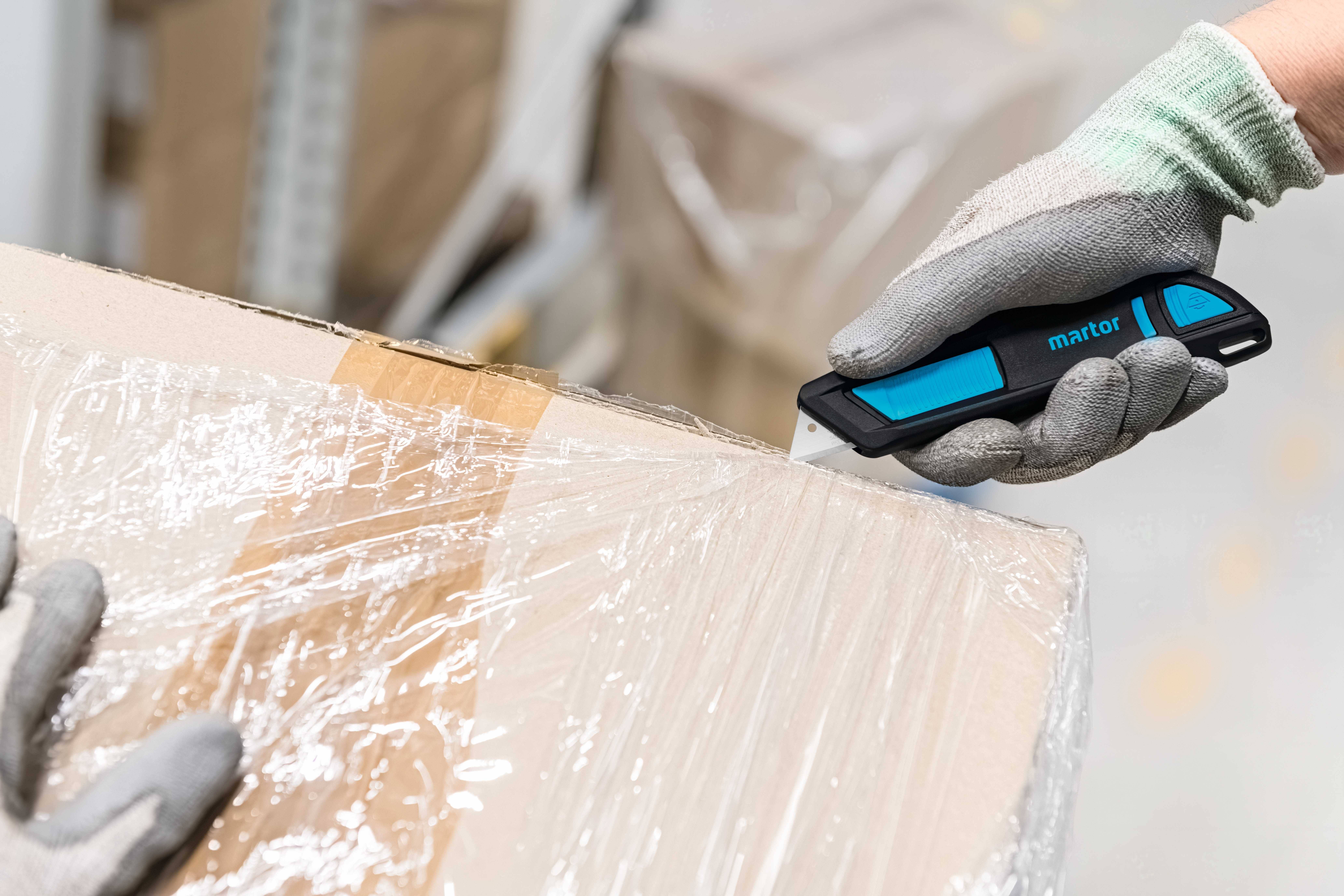 Eine Person mit Handschuhen öffnet vorsichtig einen Karton, indem sie eine Schutzfolie mit einem blauen Sicherheitsmesser durchschneidet.