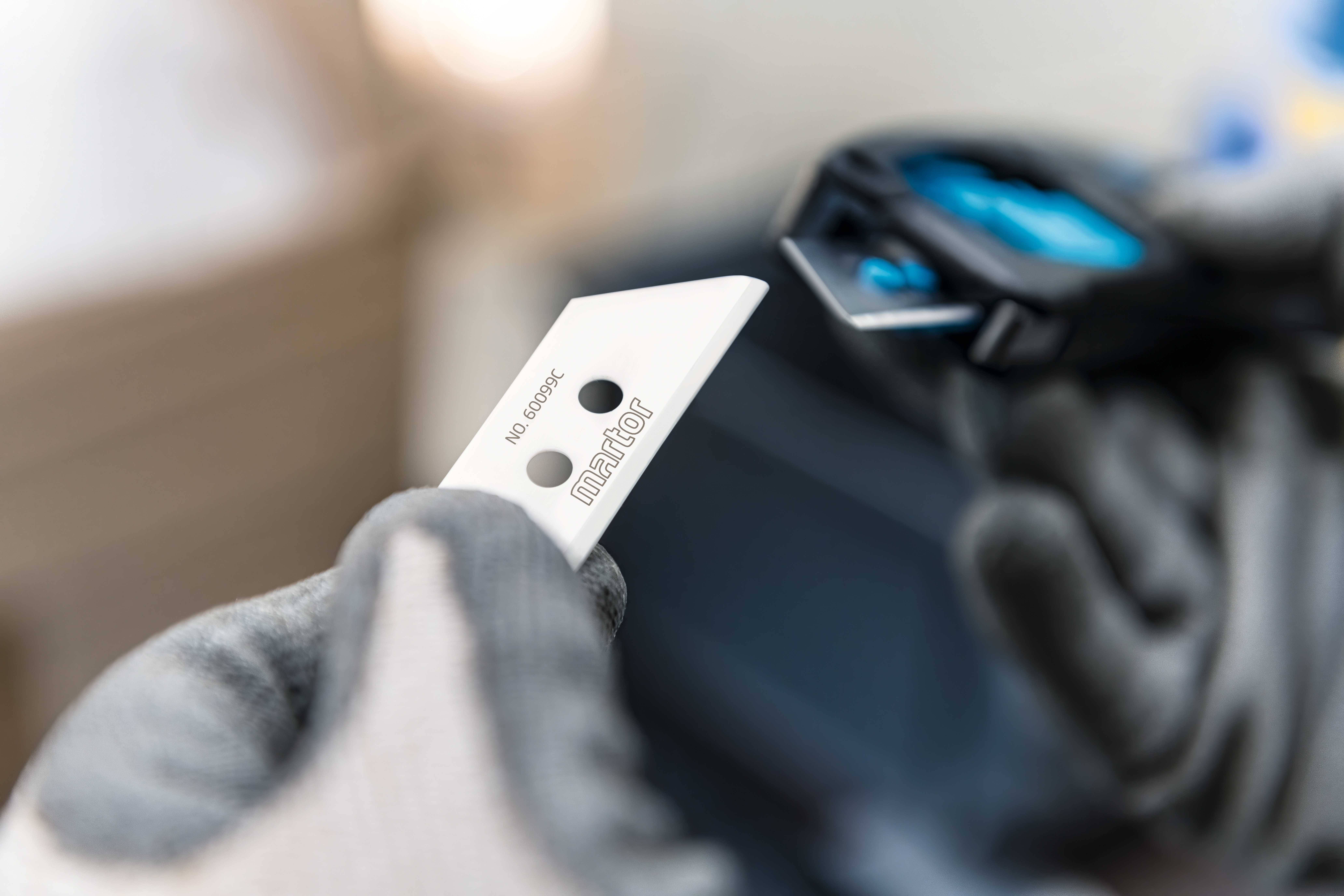 Eine Hand hält ein scharfes Messerklinge mit der Aufschrift 'martor', während die andere Hand ein blaues Werkzeug bereitstellt.