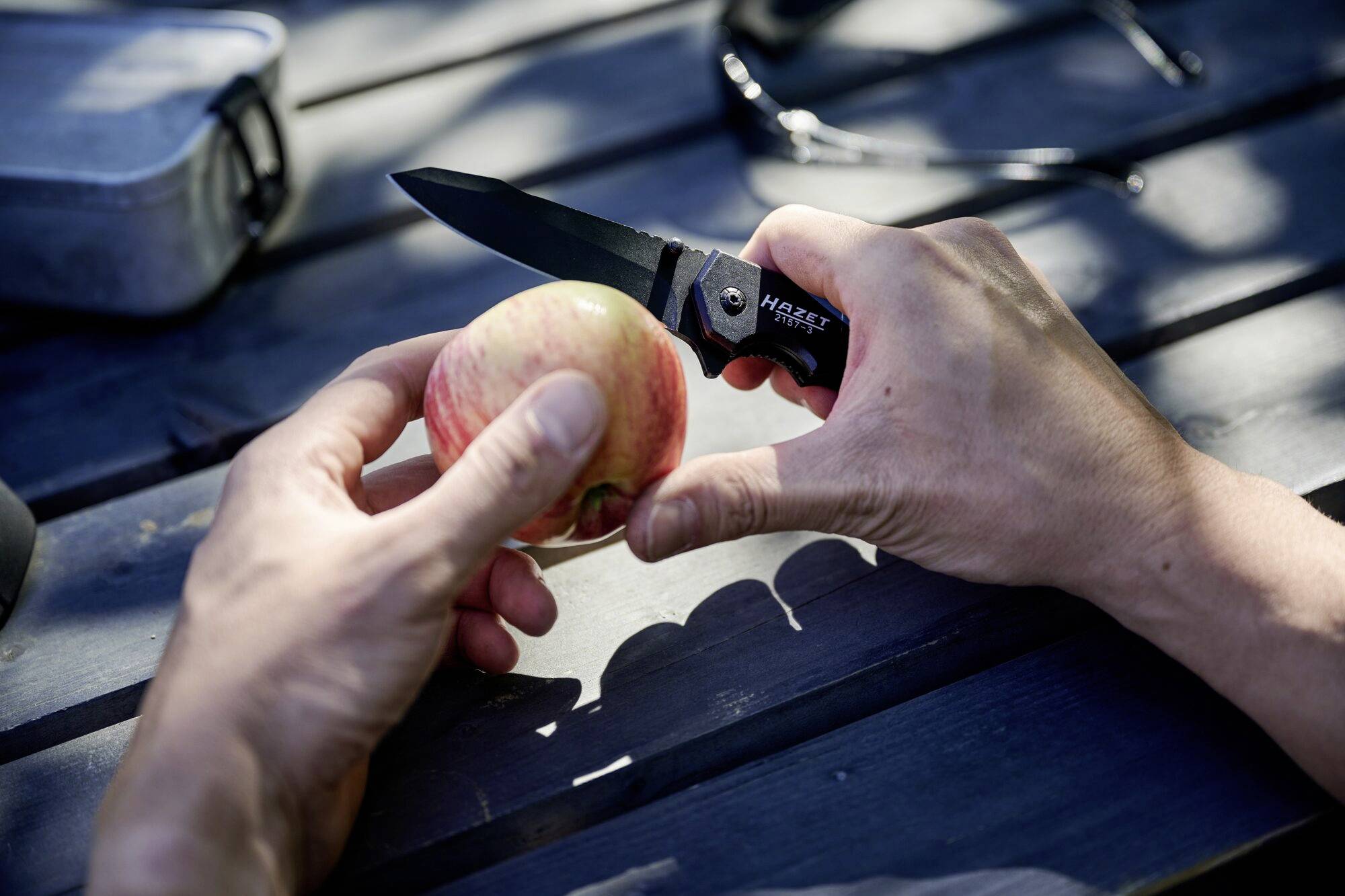 Eine Person schält einen Apfel mit einem Taschenmesser auf einem Holzpicknicktisch im Freien. Schatten fallen auf den Tisch.
