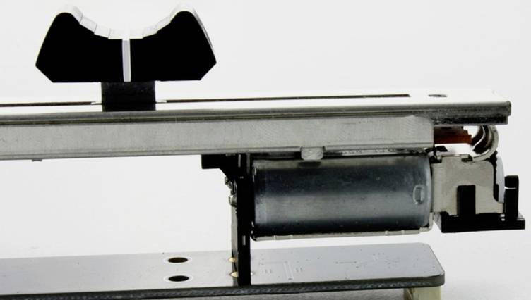 Nahaufnahme eines mechanischen Geräts mit einem Elektromotor und einem Hebelmechanismus auf einer Metallbasis vor weißem Hintergrund.
