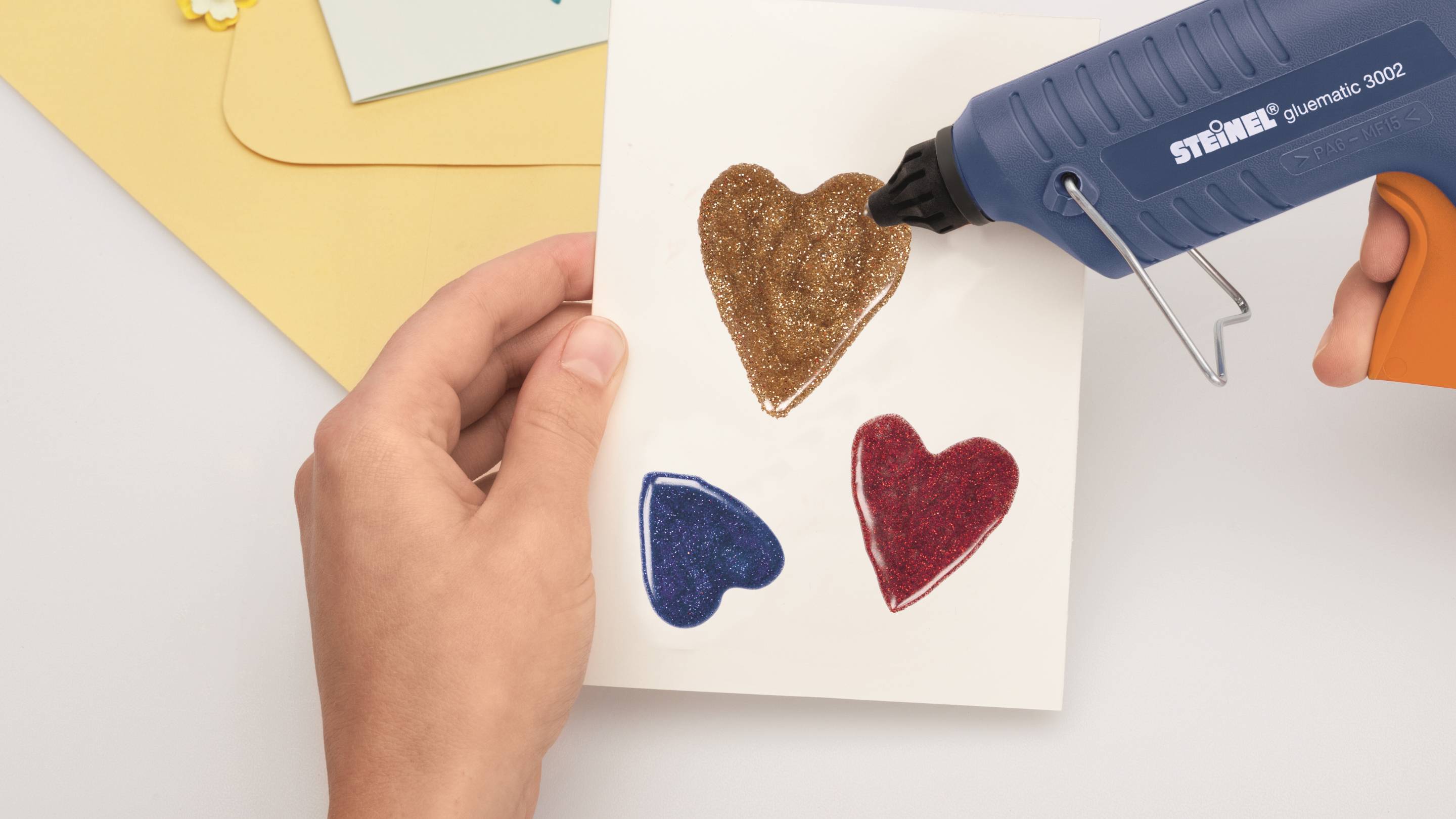 Eine Hand hält eine Karte mit drei aufgeklebten Herzen. Eine Heißklebepistole wird verwendet, um die Herzen zu befestigen.