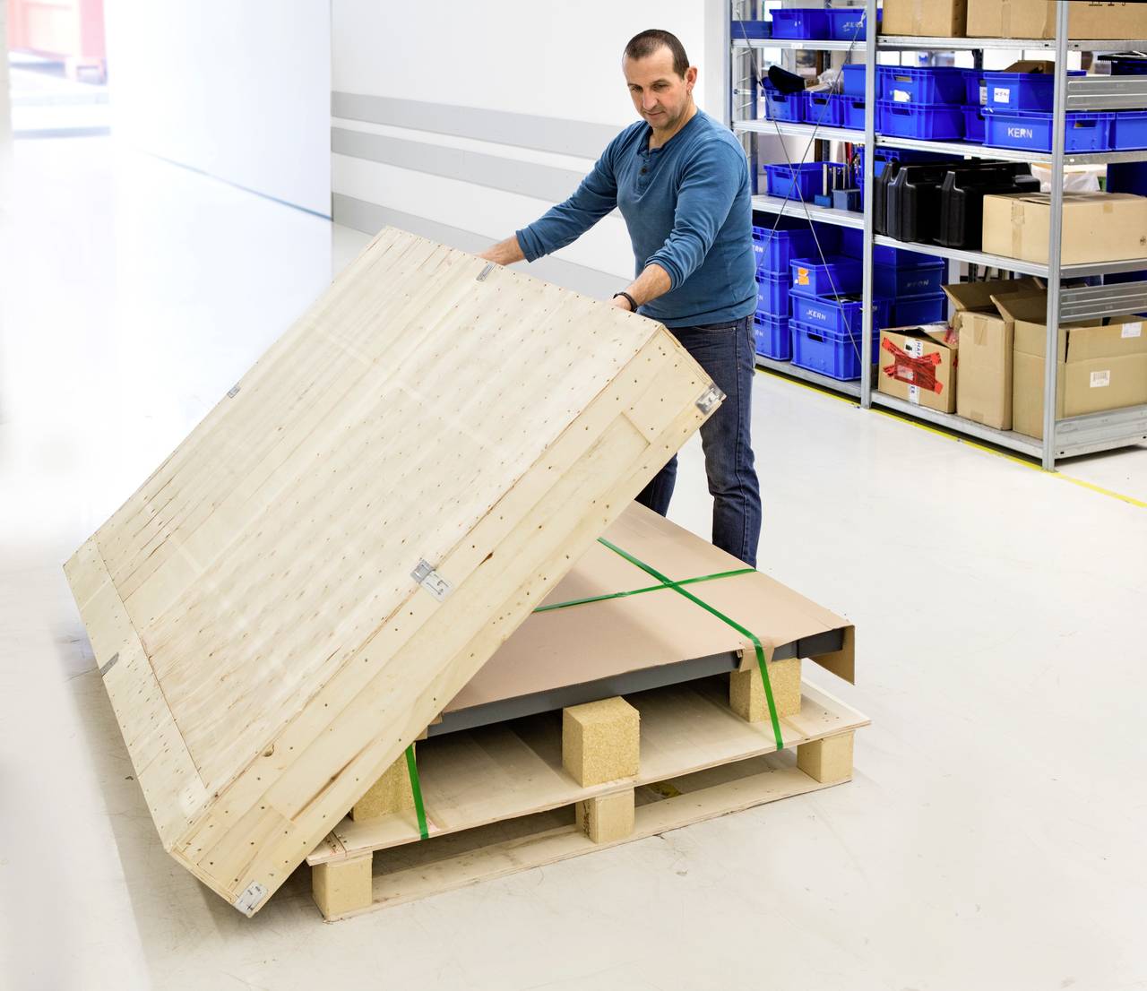 Ein Mann in einem Lager öffnet eine große Holzkiste, die auf einer Palette steht. Im Hintergrund sind Regale mit blauen Kisten.