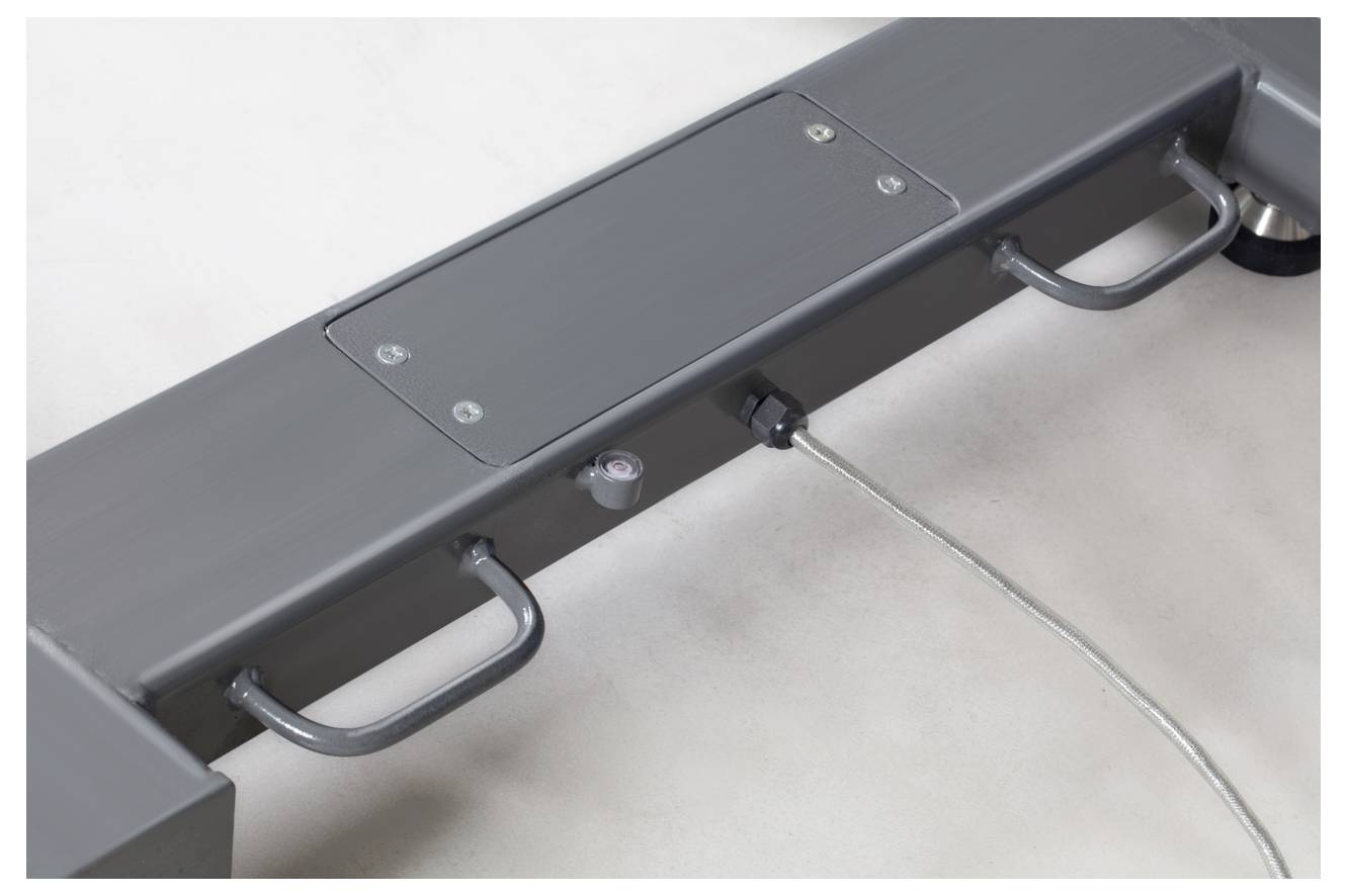 Metallisches Bauteil einer industriellen Maschine mit zwei Griffen und einem Anschlusskabel auf einer grauen Oberfläche.