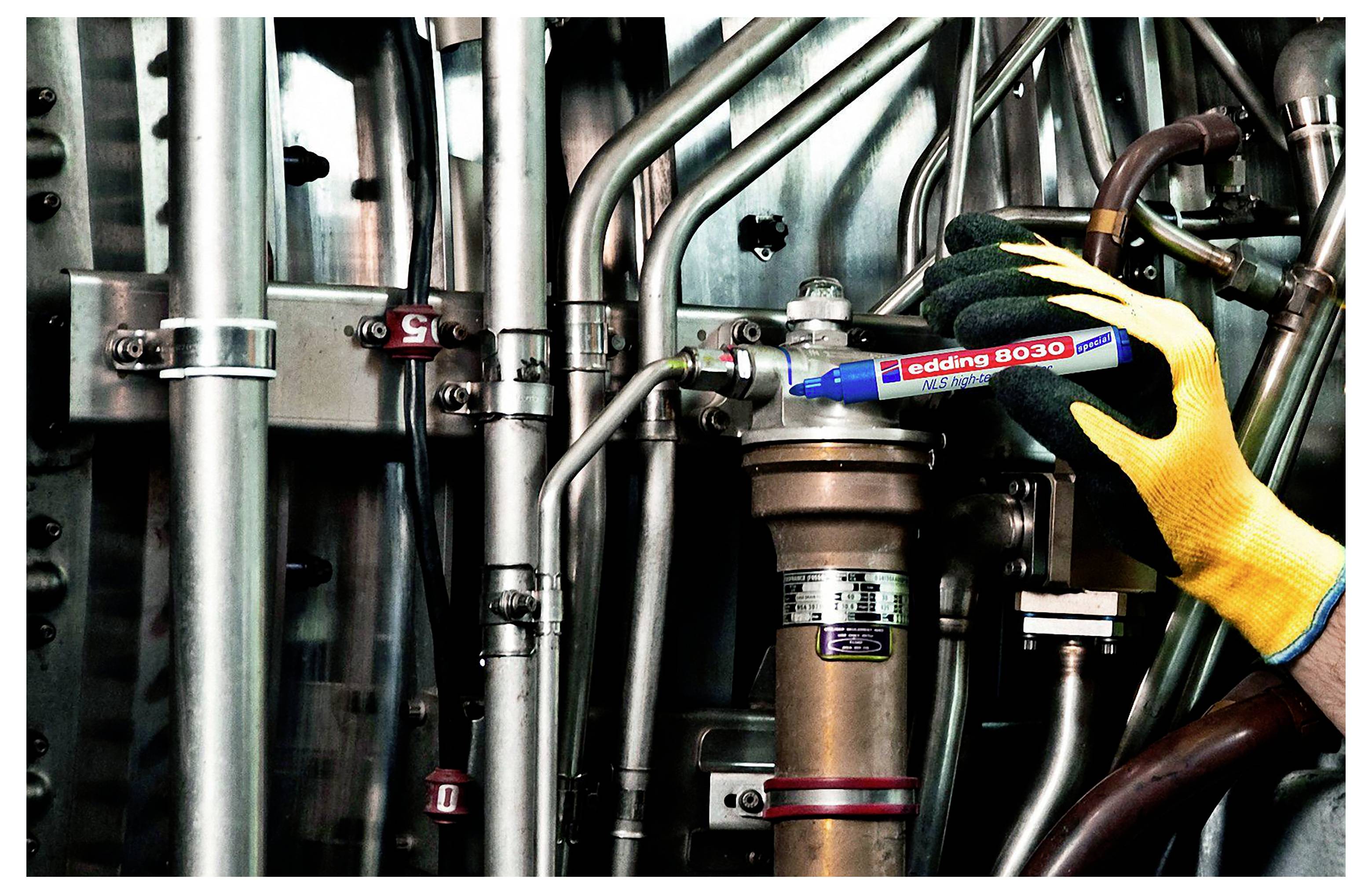 Handschuh trägt eine blaue „edding 8030"-Markierung auf einem Metallrohr, das sich zwischen industrieller Maschinerieausrüstung und Rohrleitungen befindet.