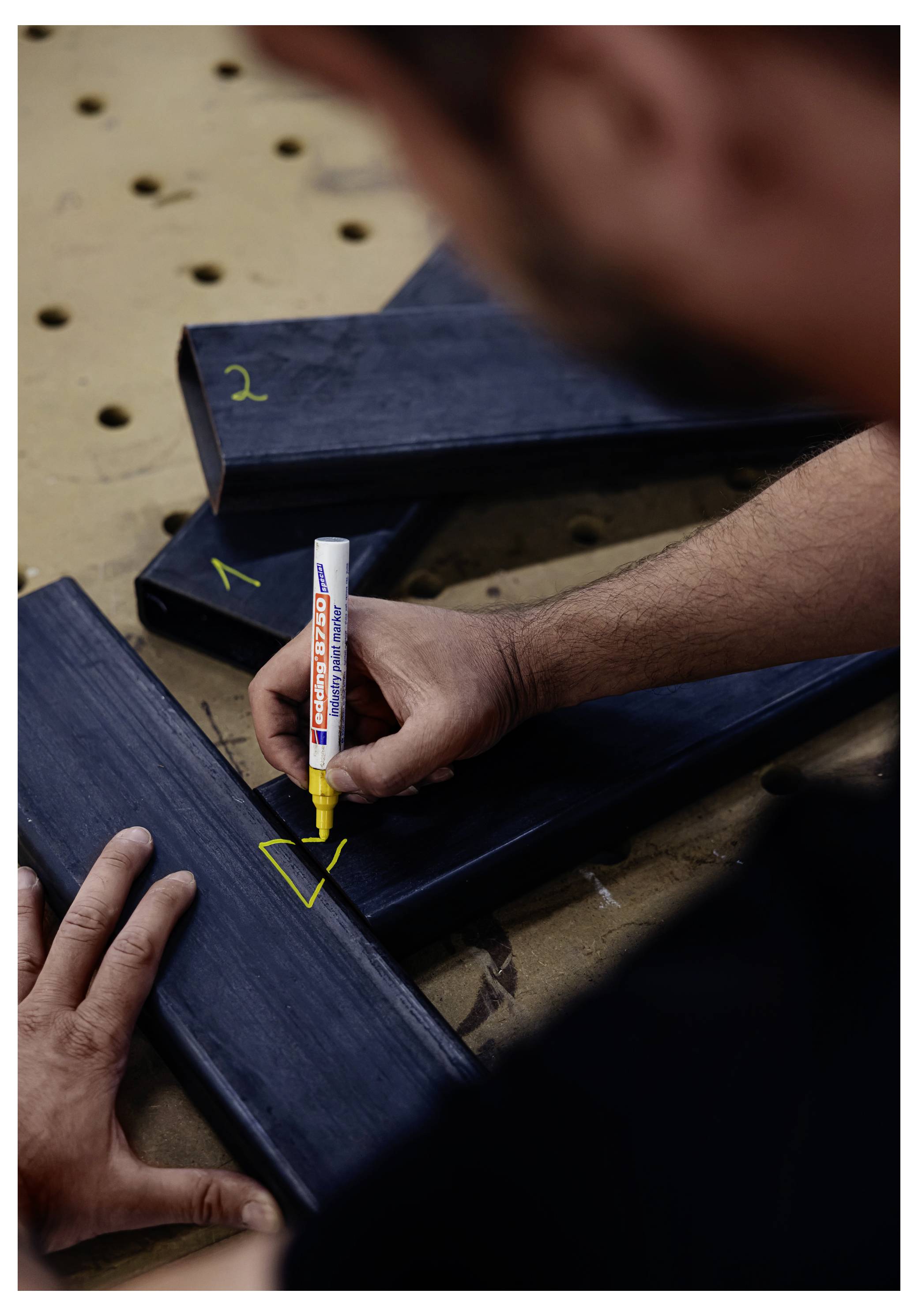 Eine Person markiert mit einem gelben Marker eine V-förmige Schnittlinie auf einem dunklen Holzrahmen oder Brett an einer Werkbank.