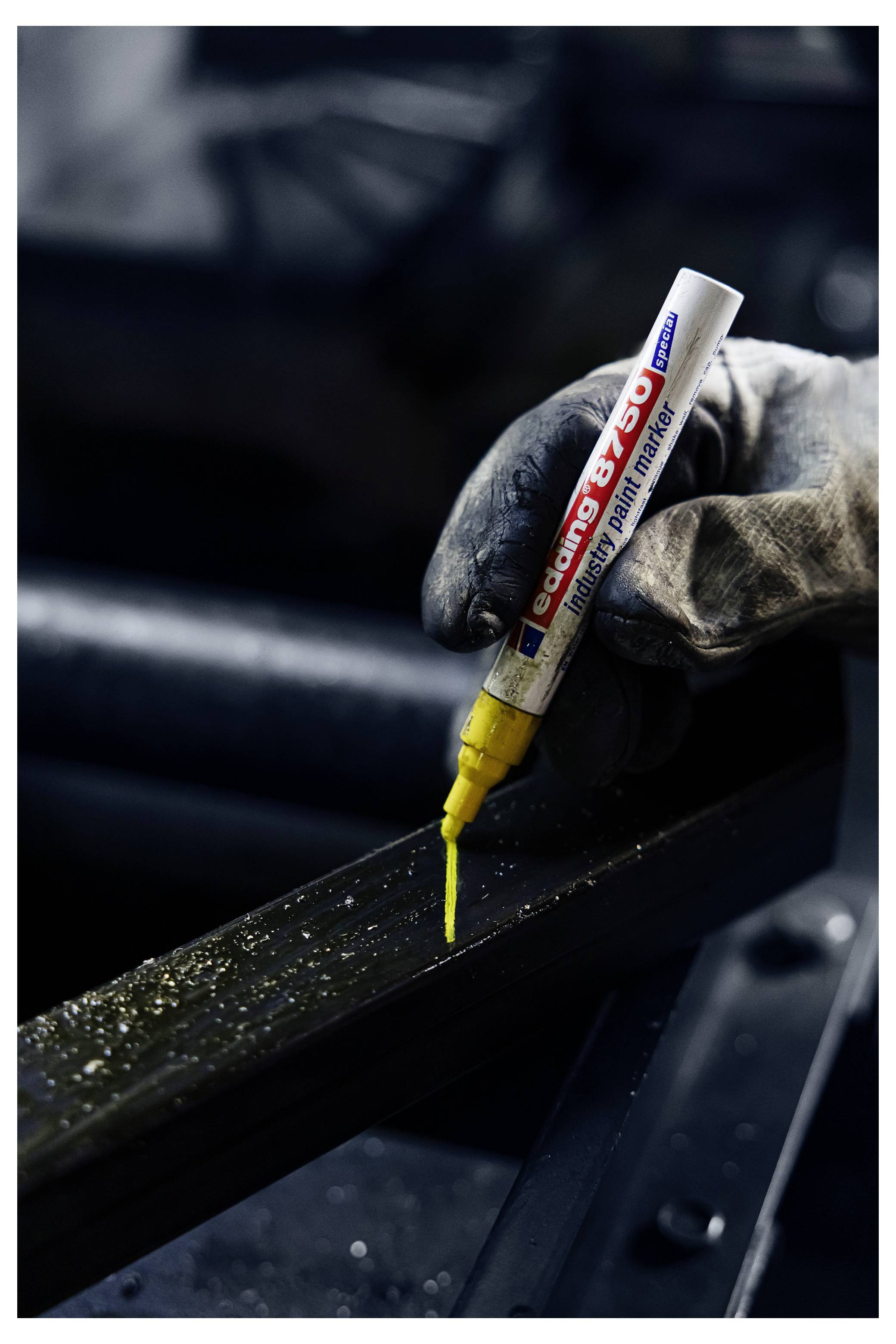 Hand mit Handschuh, die einen gelben Lackmarker verwendet, um eine Linie auf einem schwarzen Metallträger in einer Werkstatt zu zeichnen.