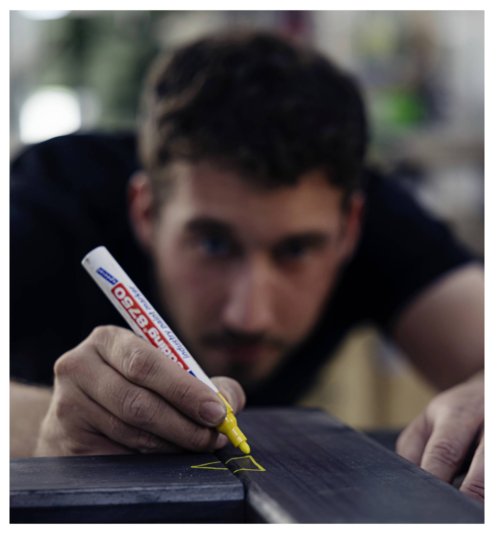 Ein Mann verwendet einen gelben Marker, um eine Linie auf einer dunklen Holzoberfläche zu zeichnen und konzentriert sich dabei intensiv auf seine Arbeit.