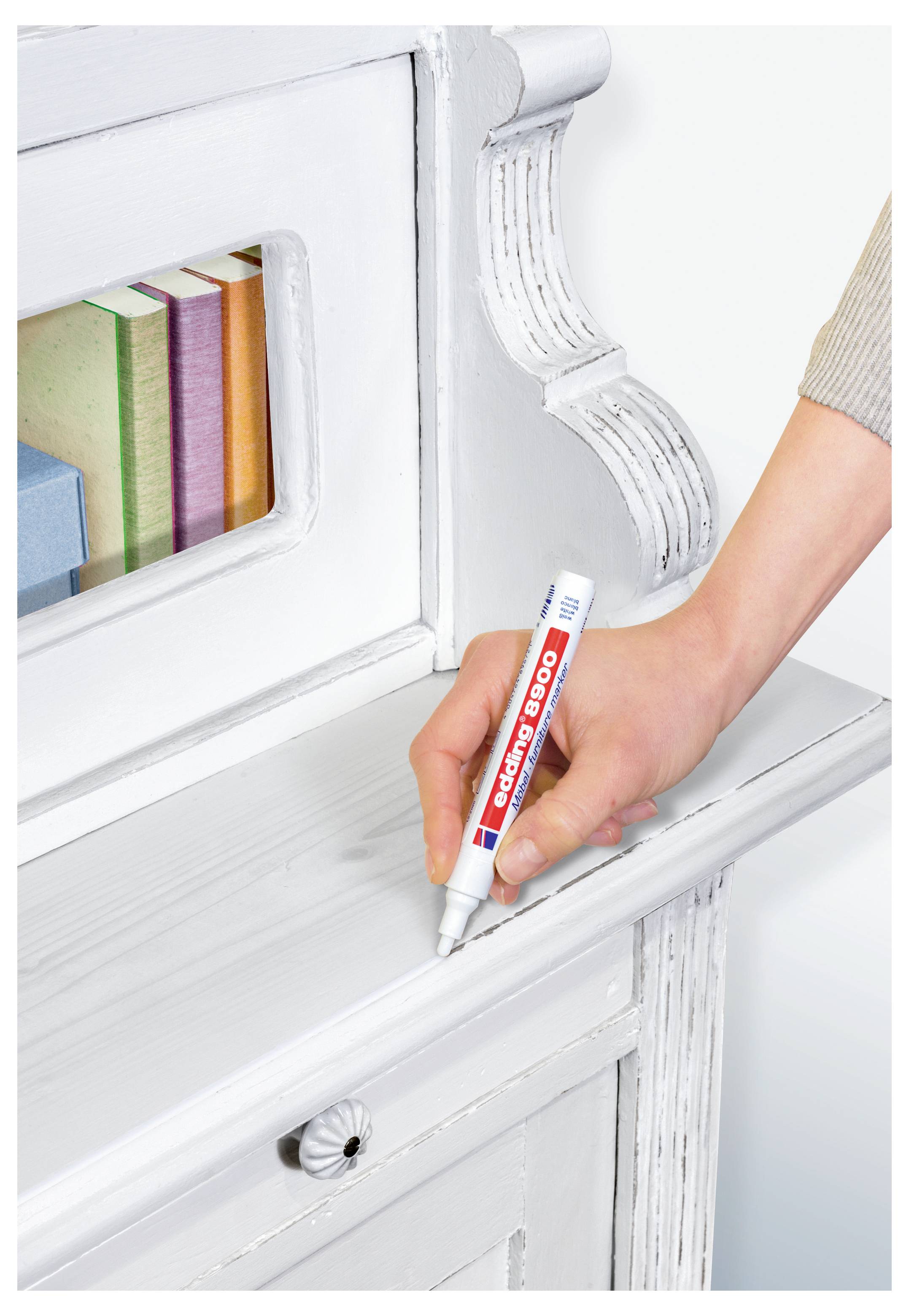 Eine Hand benutzt einen weißen Korrekturstift, um ein weiß angestrichenes Holzregal oder einen weißen Holzschrank zu retuschieren.
