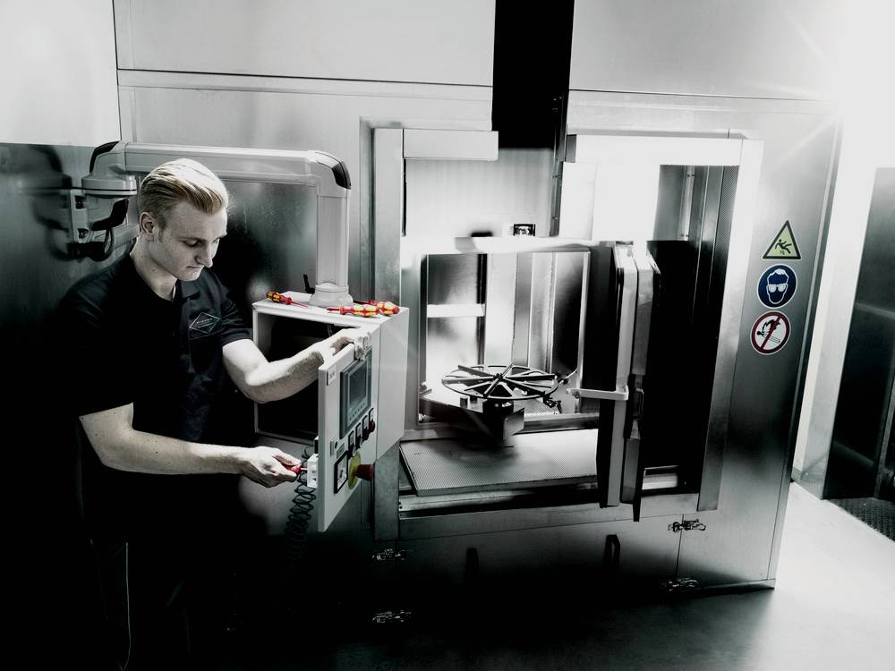 Eine Person bedient eine industrielle Maschine mit einem Schalttableau. Warn- und Sicherheitssymbole sind an der Maschine angebracht.