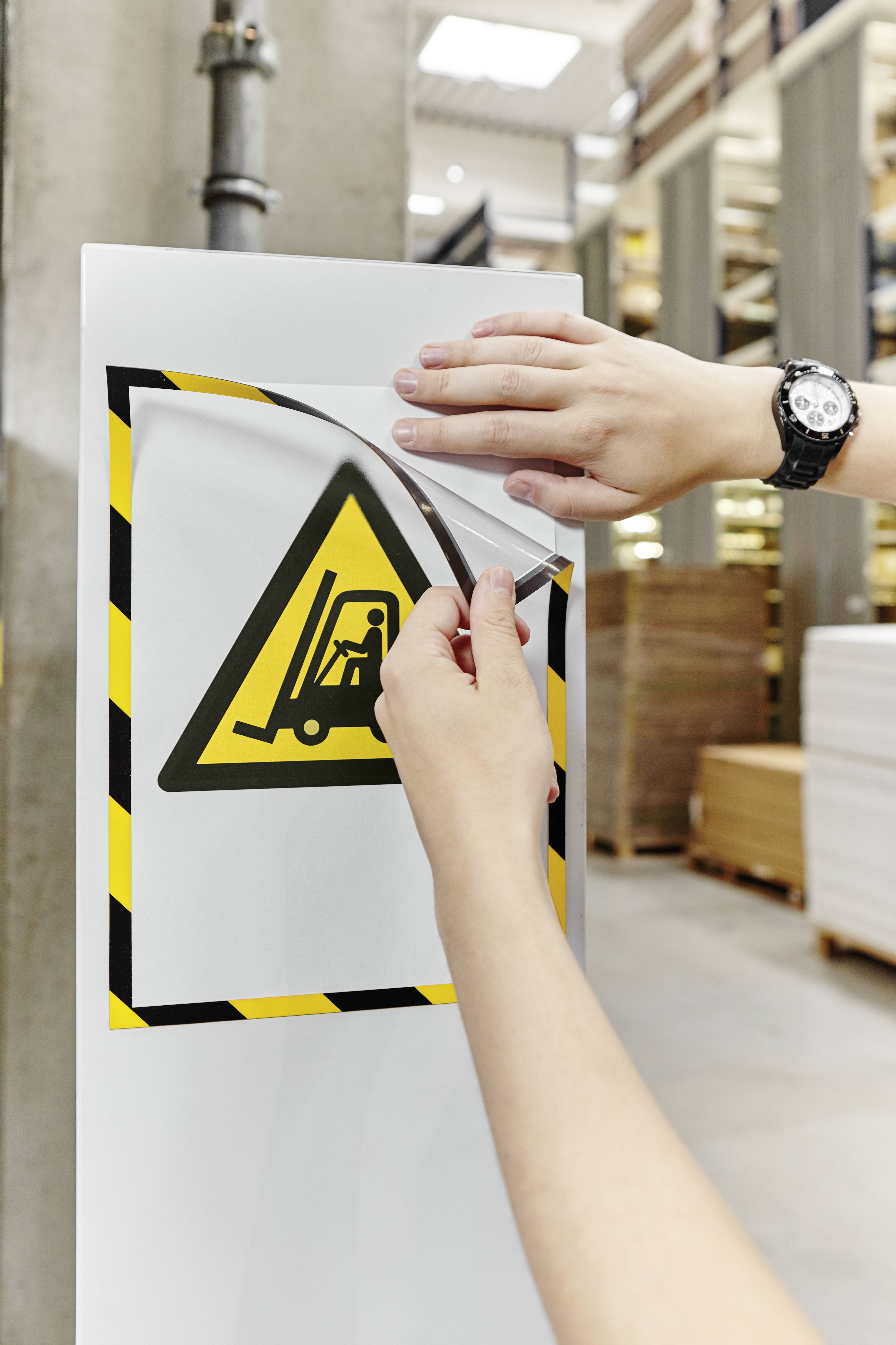 Eine Person bringt ein Warnschild 'Achtung Gabelstapler' an einer Wand in einem Lager an. Im Hintergrund sind Regalreihen sichtbar.