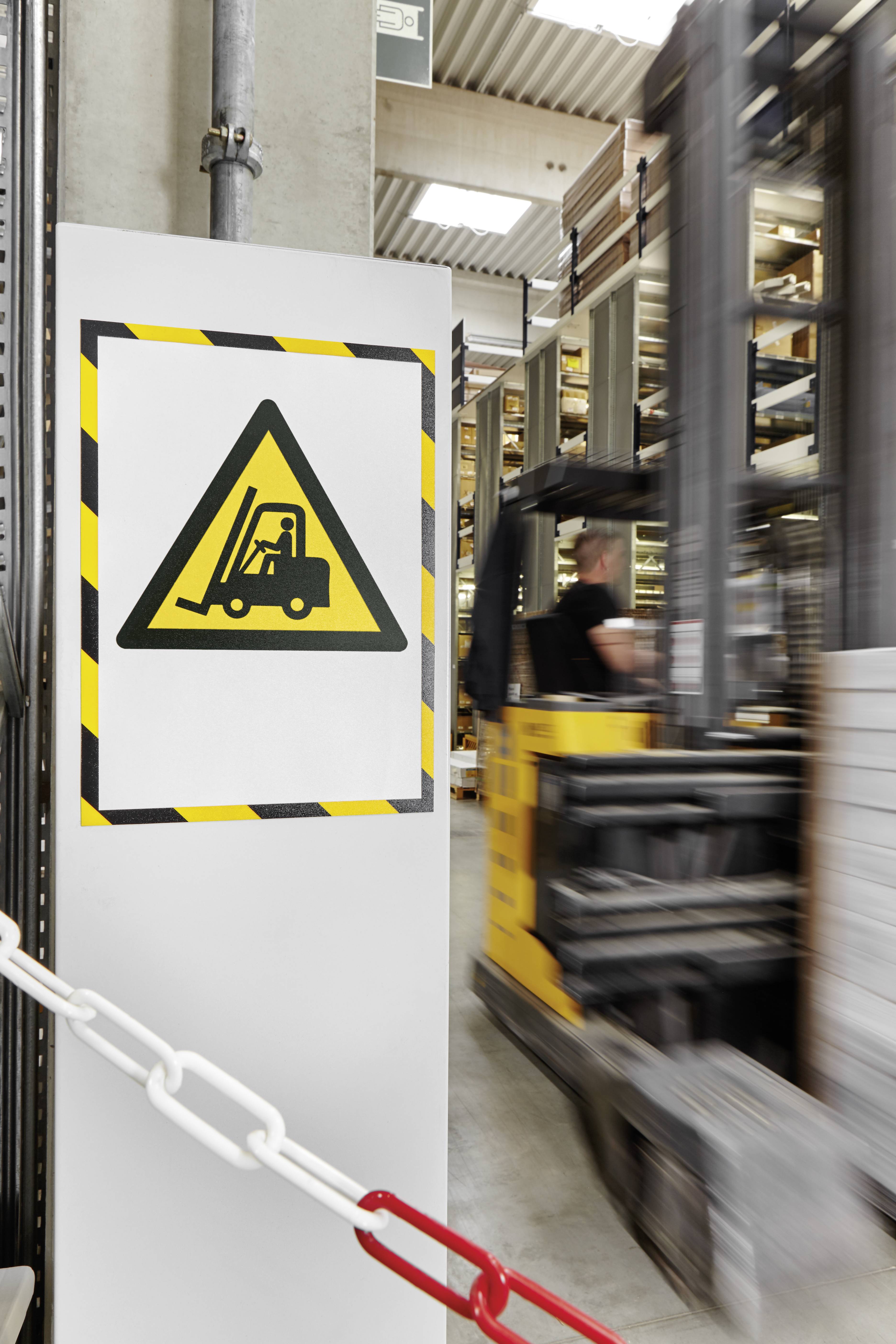 Ein Schild mit einem Gabelstaplersymbol warnt vor einer Gabelstaplerzone in einem Industriegebäude. Im Hintergrund ein fahrender Gabelstapler.