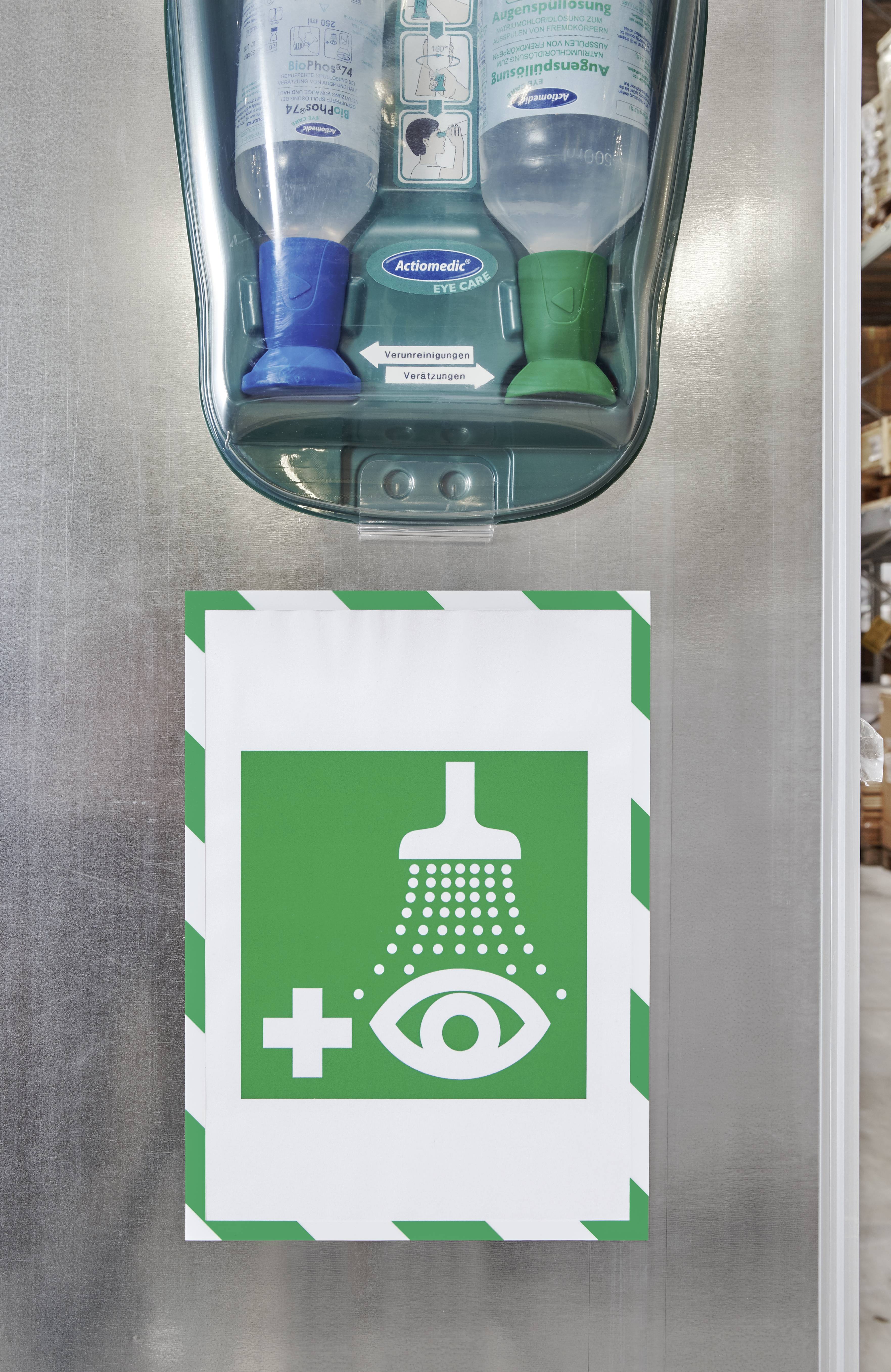 Notfall-Augenspülstation an einer Wand mit grünem und weißem Schild mit Augenduschen-Symbol, darunter zwei Flaschen Augenspülflüssigkeit.