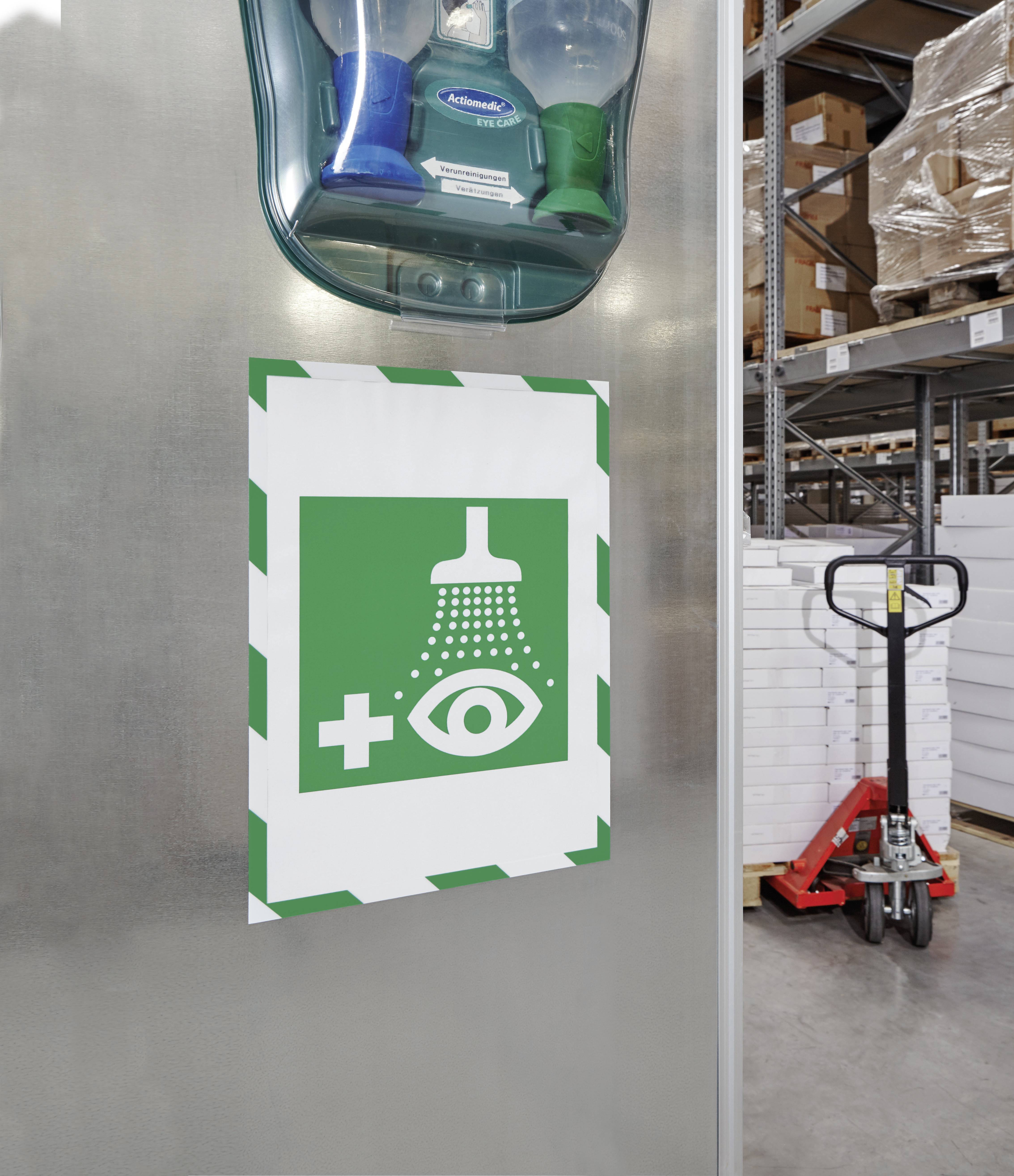 Ein grünes Symbol für eine Augendusche ist an der Wand eines Lagers befestigt, nahe einem Stapel weißer Kisten und einem Gabelhubwagen.