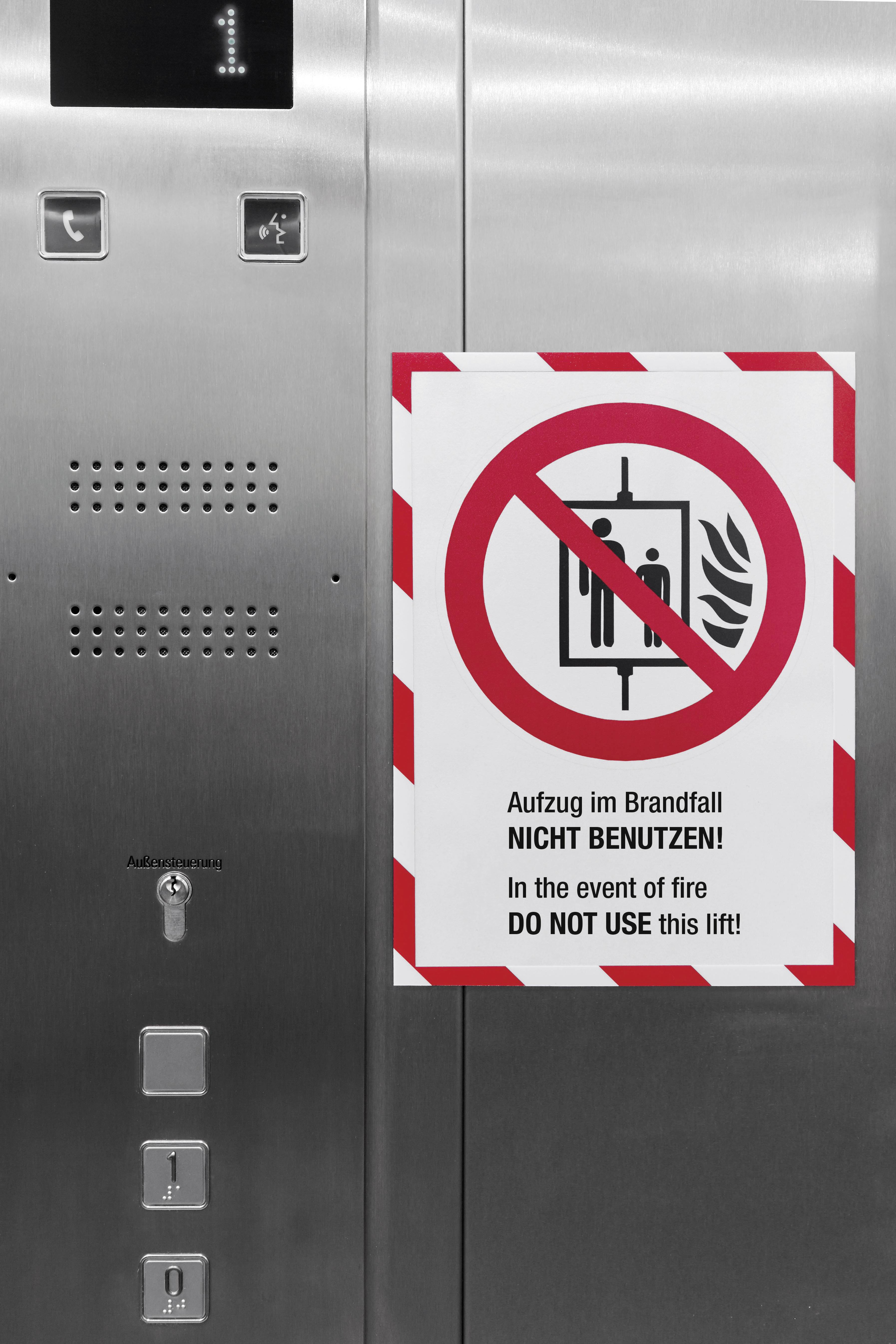 Das Bild zeigt eine Aufzugstür mit einem Warnschild: 'Aufzug im Brandfall NICHT BENUTZEN!'. Ein durchgestrichenes Symbol begleitet den Text.