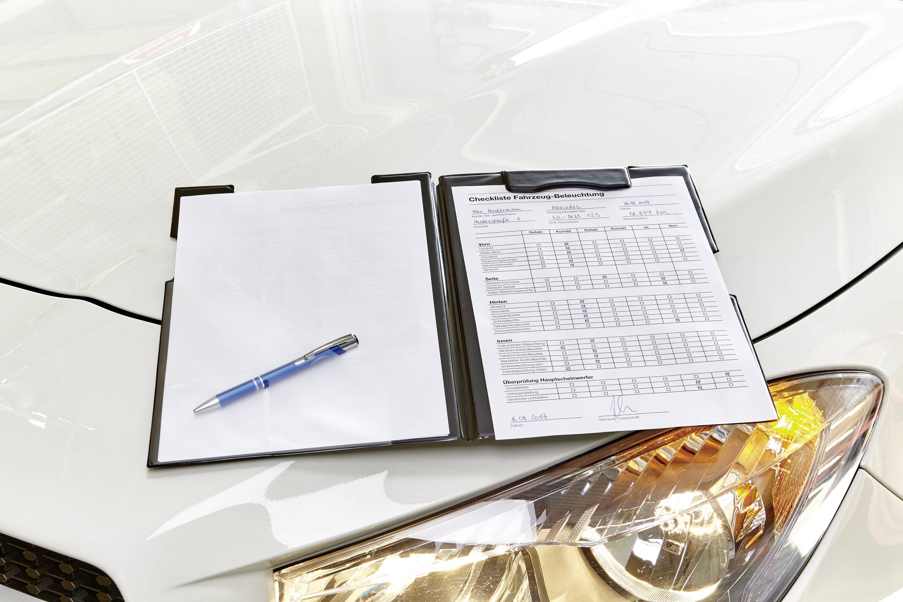 Ein Klemmbrett mit Formular und Stift liegt auf einer Motorhaube eines Autos. Das Formular enthält Tabellen und handschriftliche Notizen.