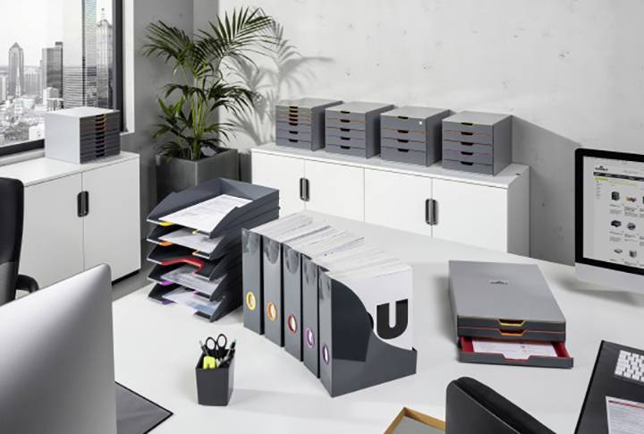 Ein modernes Büro mit weißen Möbeln, mehreren Aktenordnern und Ablagen auf dem Schreibtisch. Im Hintergrund Stadtblick aus dem Fenster.