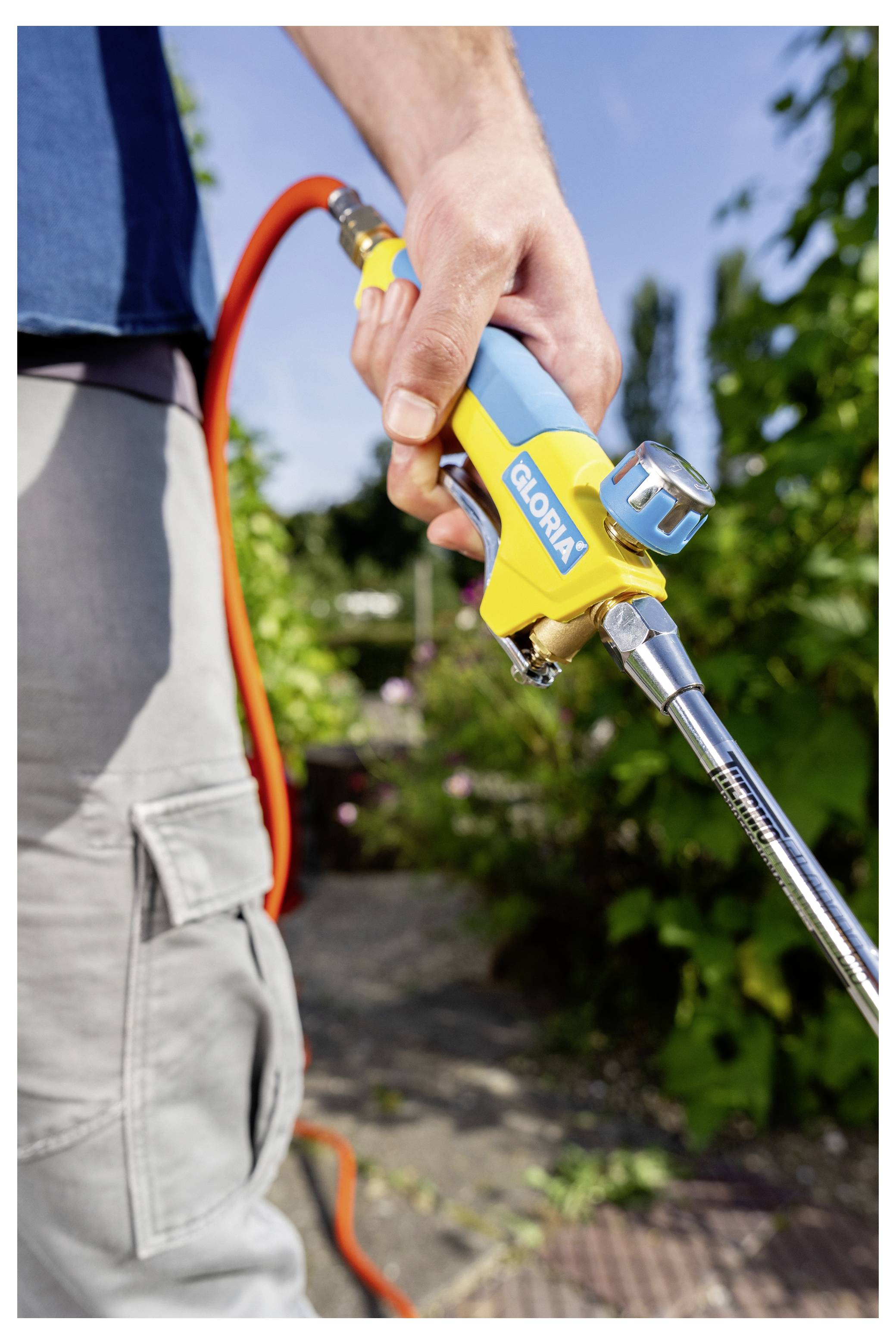 Nahaufnahme einer Person, die draußen eine gelbe GLORIA-Gartenspritzpistole hält, die an einem orangefarbenen Schlauch befestigt ist, und diese nach unten gerichtet zum Bewässern oder Reinigen verwendet.