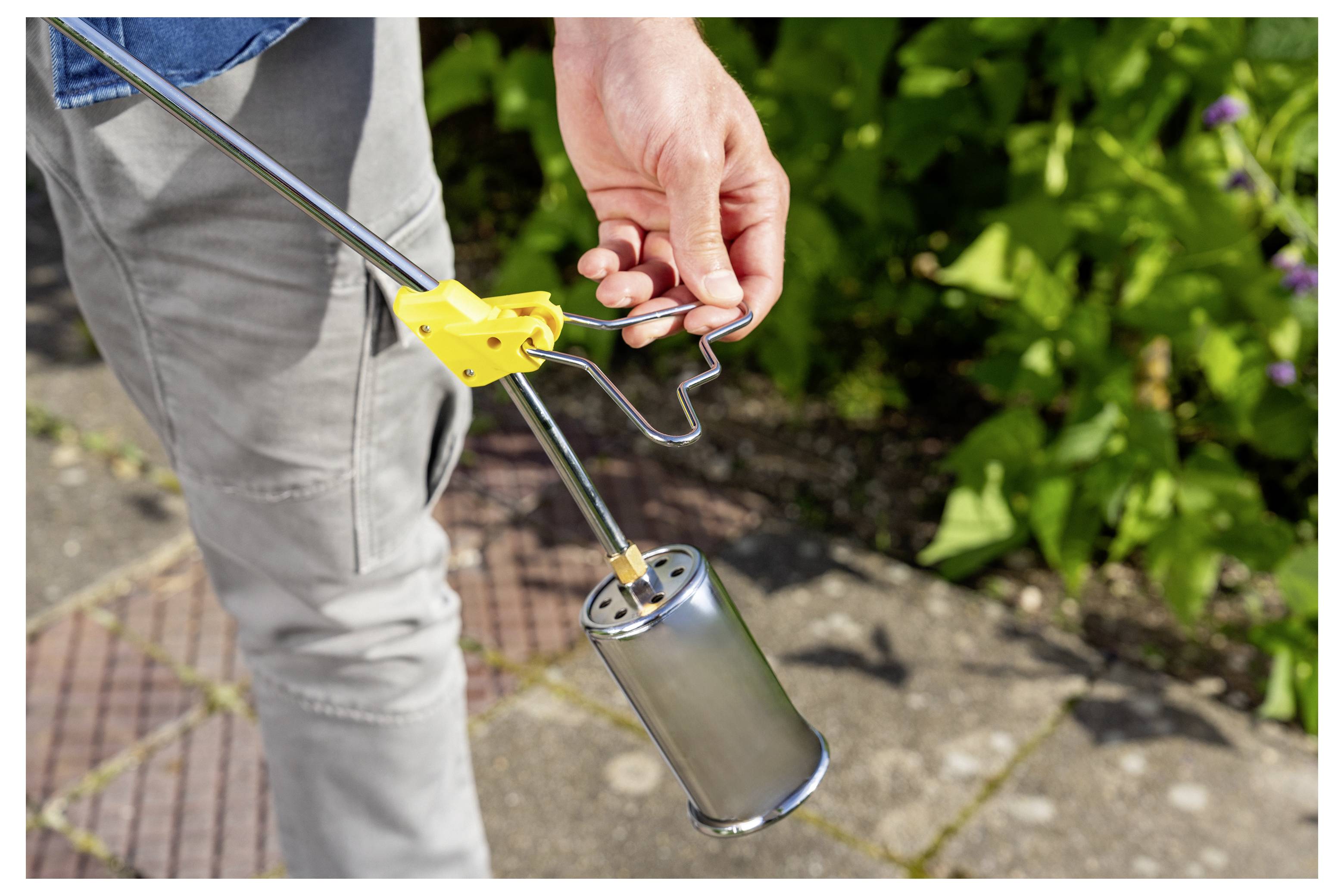Person, die eine tragbare Gartenfackel mit einem gelben Zündgriff und einer Metallbüchse hält, im Freien auf einem gepflasterten Weg in der Nähe von Grünflächen.