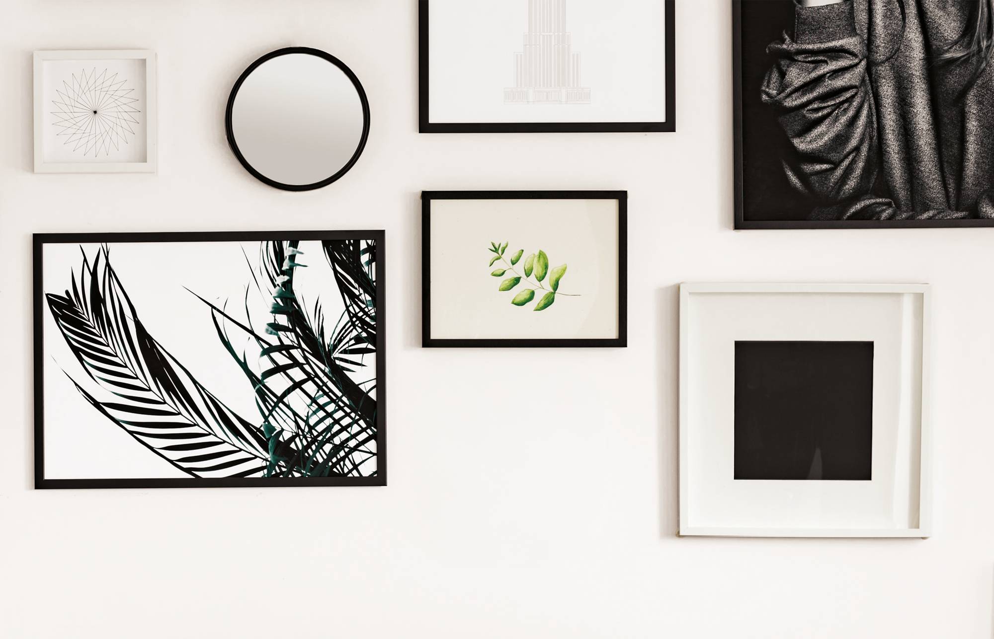 Galeriewand mit gerahmten Kunstwerken und Drucken, einschließlich schwarz-weißer botanischer Designs, einem runden Spiegel und abstrakten Bildern an einer weißen Wand.