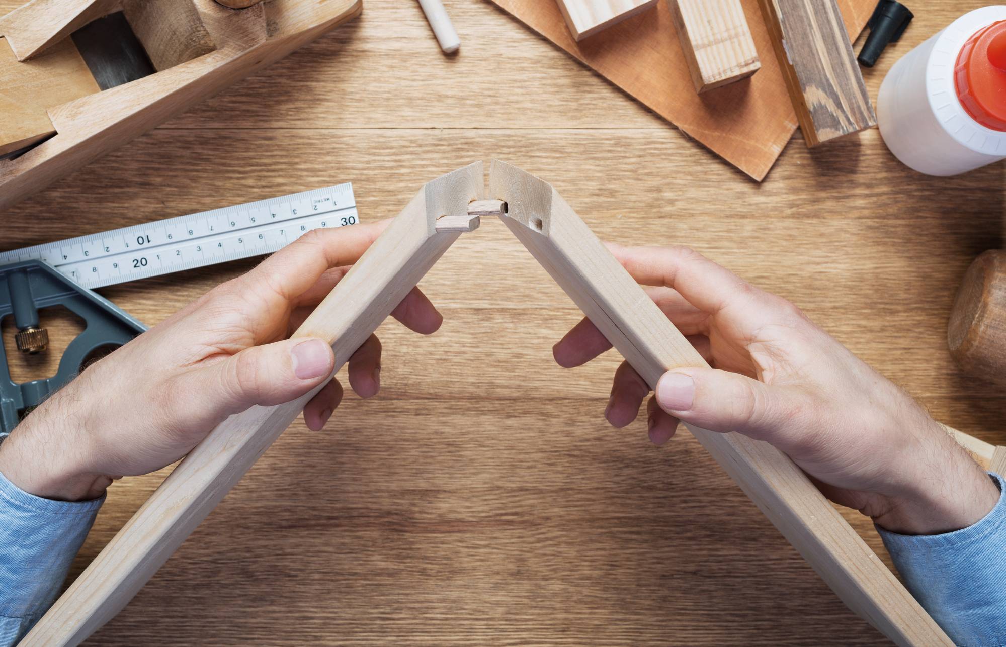 Hände, die zwei Holzrahmenstücke auf einer Werkbank zusammenbiegen und eine Ecke mit Klebstoff oder einem Verbinder in einem Holzbearbeitungsprojekt verbinden.