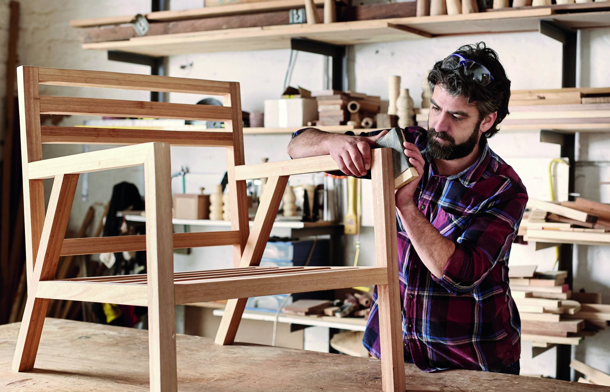 Ein Mann in einer Werkstatt montiert ein Holzstuhlgestell und bearbeitet die Sitzkante mit einem Handwerkzeug.