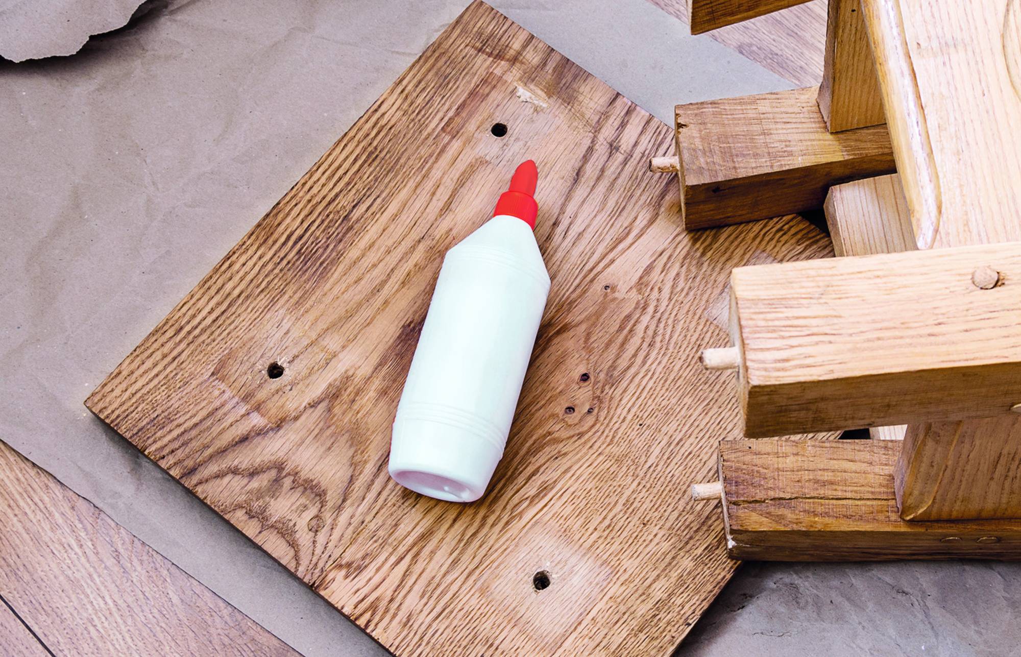 Flasche mit weißem Kleber und roter Düse auf einem Holzbrett neben gestapelten Holzblöcken.