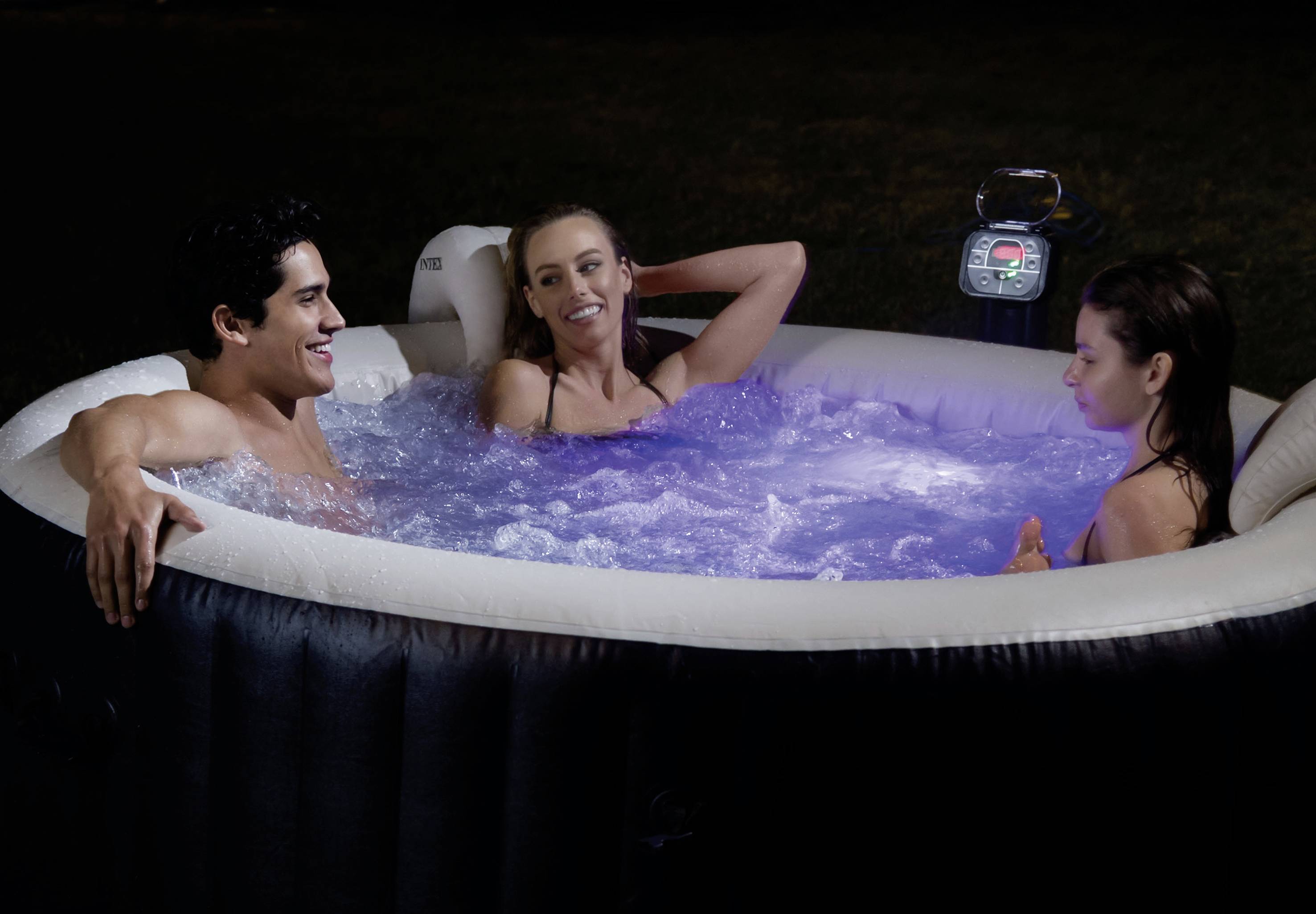 Drei Personen entspannen sich abends in einem beleuchteten aufblasbaren Whirlpool im Freien. Sie lachen und plaudern miteinander.