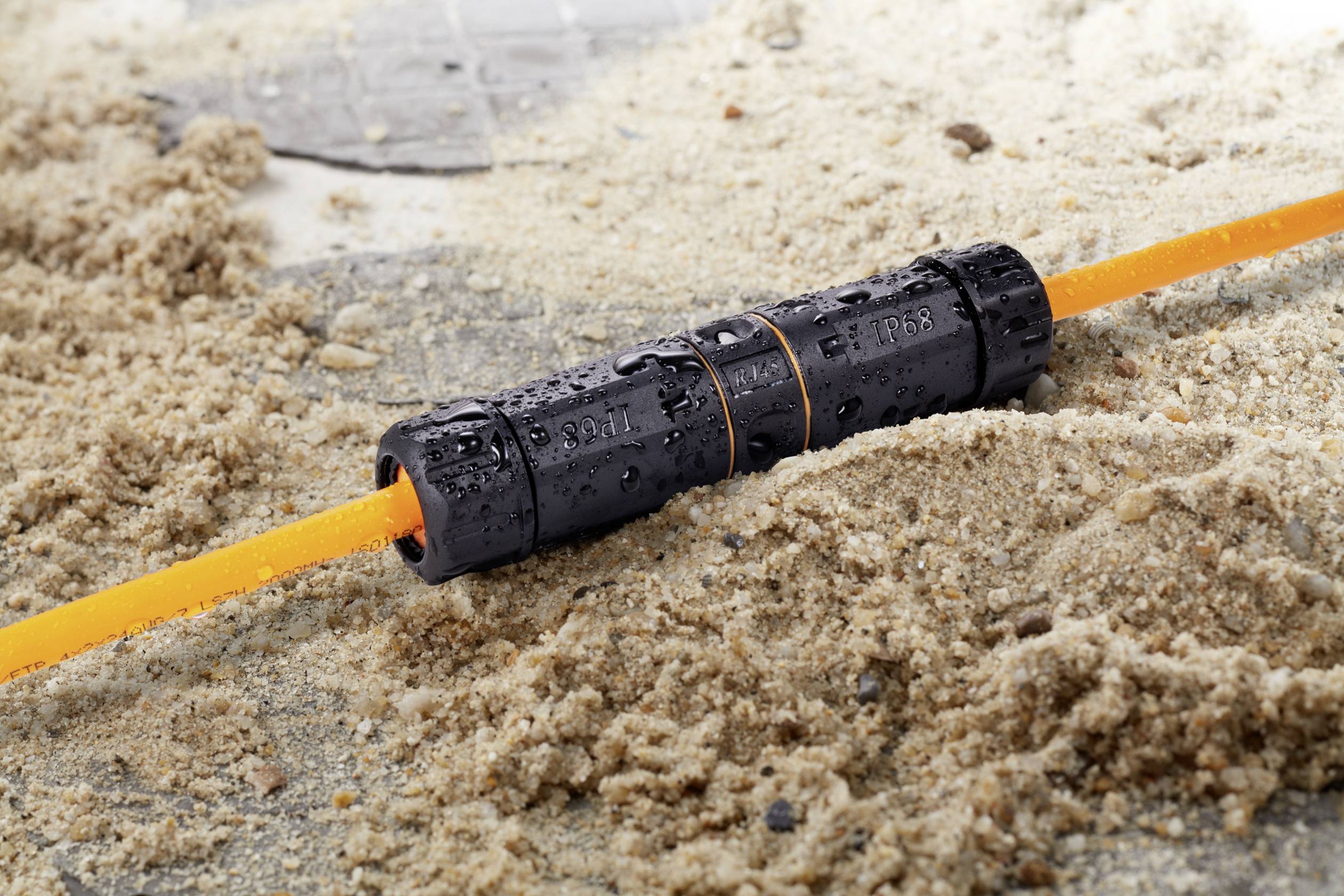 Ein wasserdichter schwarzer Verbinder mit einer orangefarbenen Leitung liegt auf sandigem Untergrund.