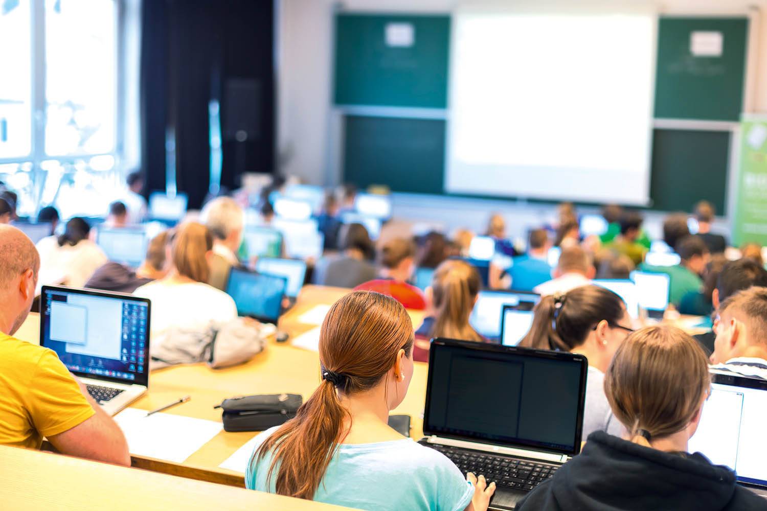 Studenten sitzen in einem Hörsaal, arbeiten auf Laptops, und konzentrieren sich auf eine Präsentation an einer Tafel.