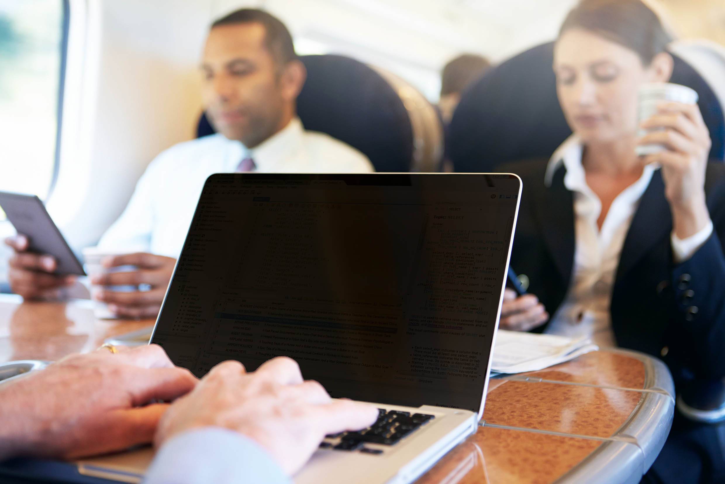 Ein Mann arbeitet an einem Laptop in einem Zugabteil, während zwei weitere Personen im Hintergrund mit einem Tablet und einem Getränk beschäftigt sind.