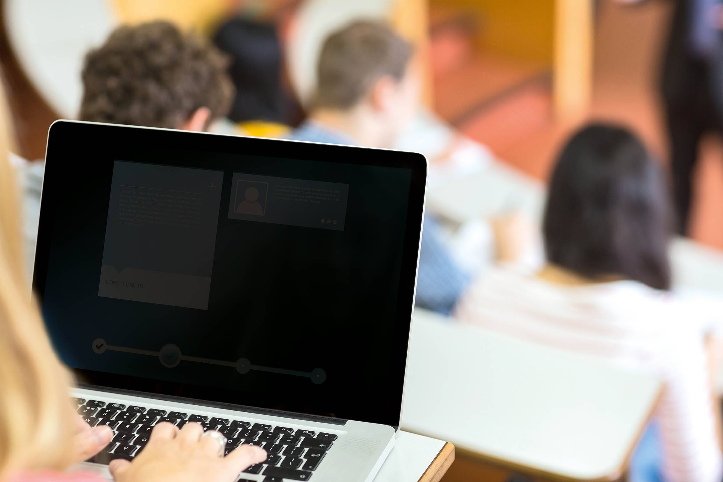 Eine Person tippt auf einem Laptop in einem Klassenzimmer. Im Hintergrund sind unscharfe Studenten zu sehen, die einem Vortrag folgen.