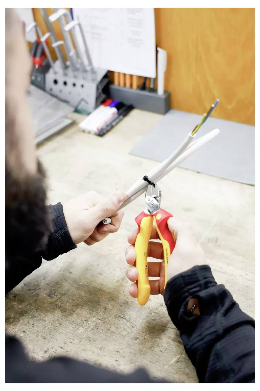 Eine Person benutzt eine Drahtschere, um ein Kabel auf einer Werkbank zu abisolieren. Verschiedene Werkzeuge und Stifte befinden sich im Hintergrund.