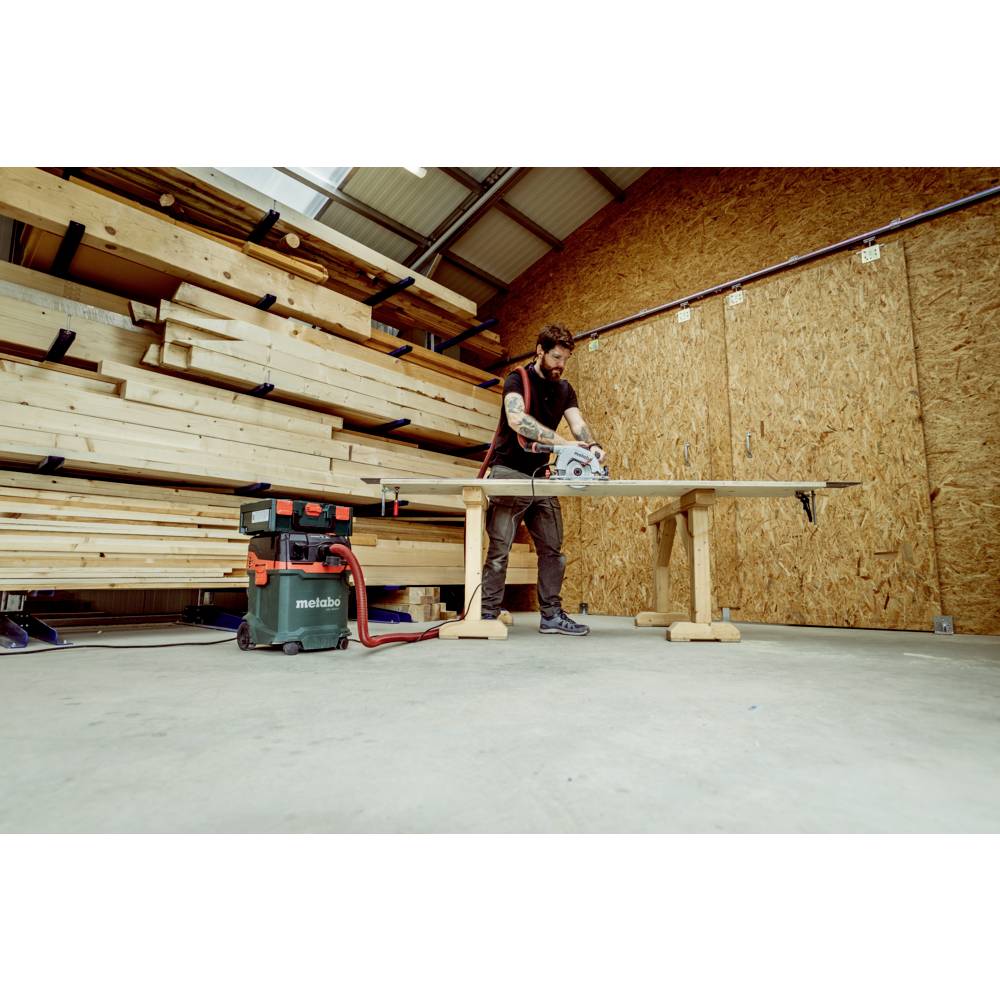 Ein Mann in einer Werkstatt schneidet eine Holzplanke auf einem Tisch mit einer Kreissäge. Im Hintergrund sind Stapel von Holz zu sehen.