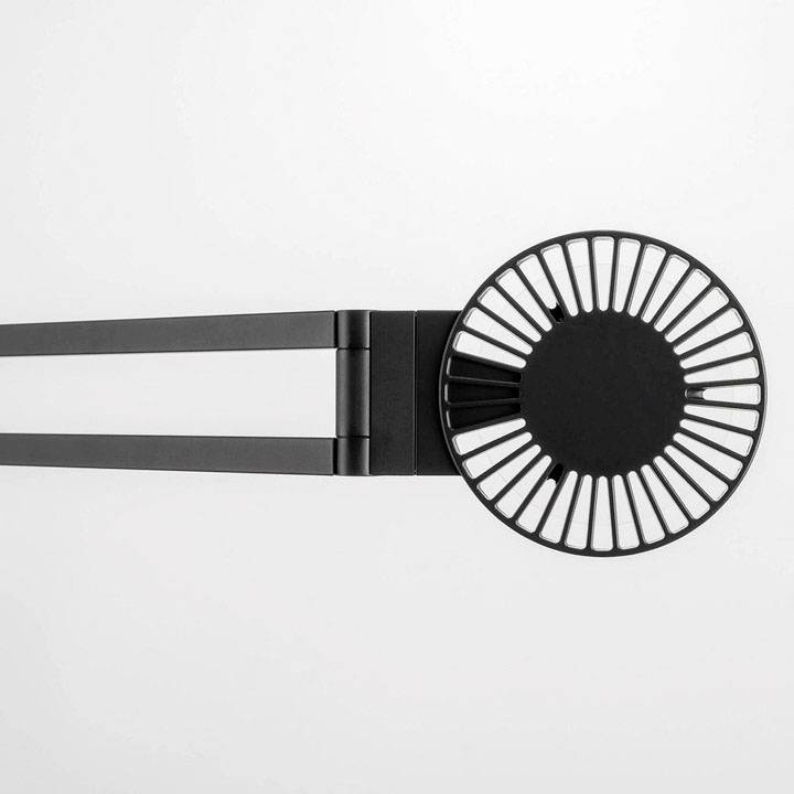 Schwarzer, runder Tischventilator mit Lamellen von oben, an einem schwarzen Arm befestigt, auf grauem Hintergrund.
