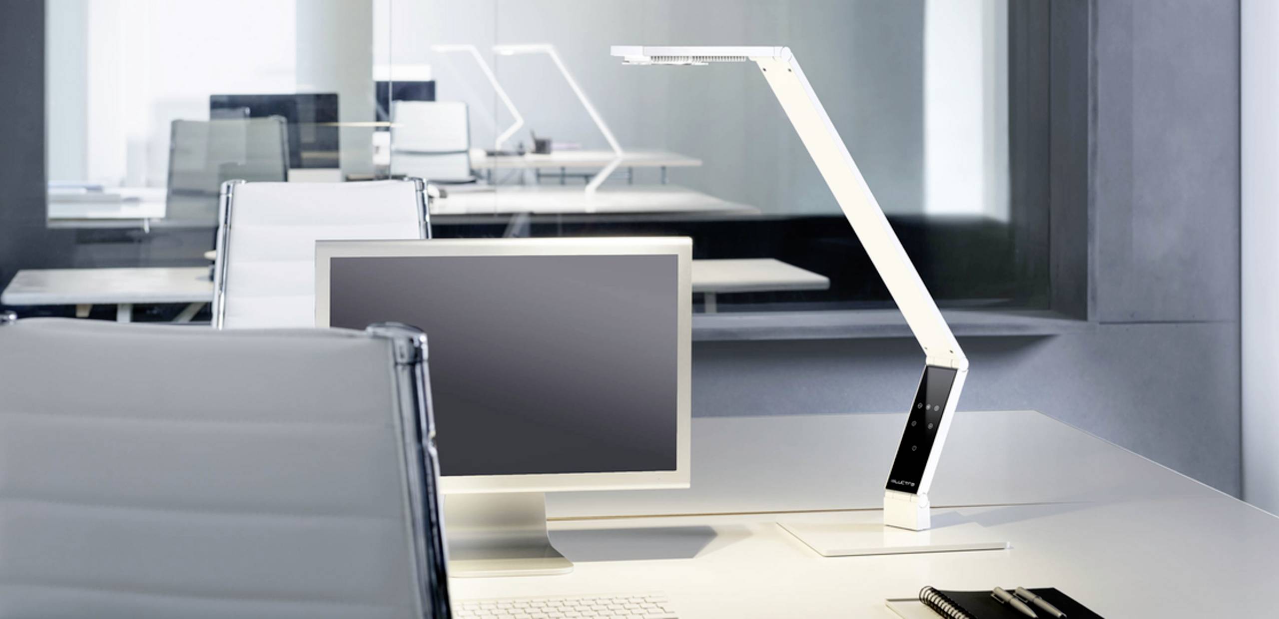 Ein moderner Schreibtisch mit einem Computerbildschirm und einer eleganten, weißen Tischlampe in einem minimalistischen Büro.