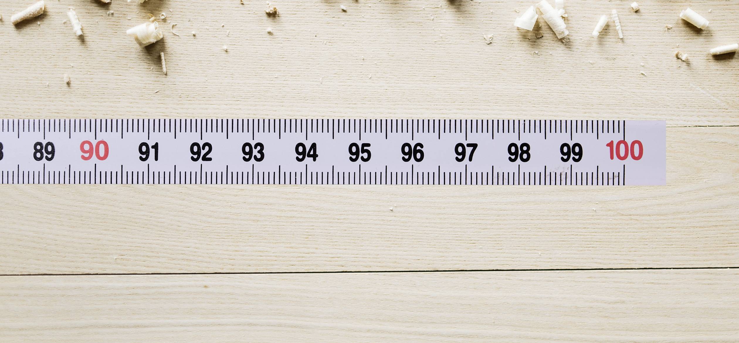Ein Maßband auf einem Holztisch zeigt Zentimeterzahlen von 90 bis 100. Holzspäne liegen um das Maßband verteilt.