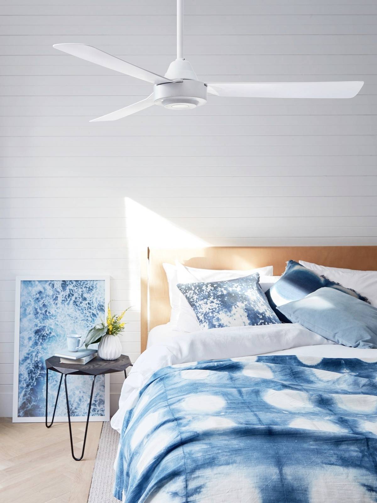 Ein modernes Schlafzimmer mit einem weißen Deckenventilator, einem Bett mit blauer Bettwäsche und Kunstwerk mit Meeresmotiv im Hintergrund.