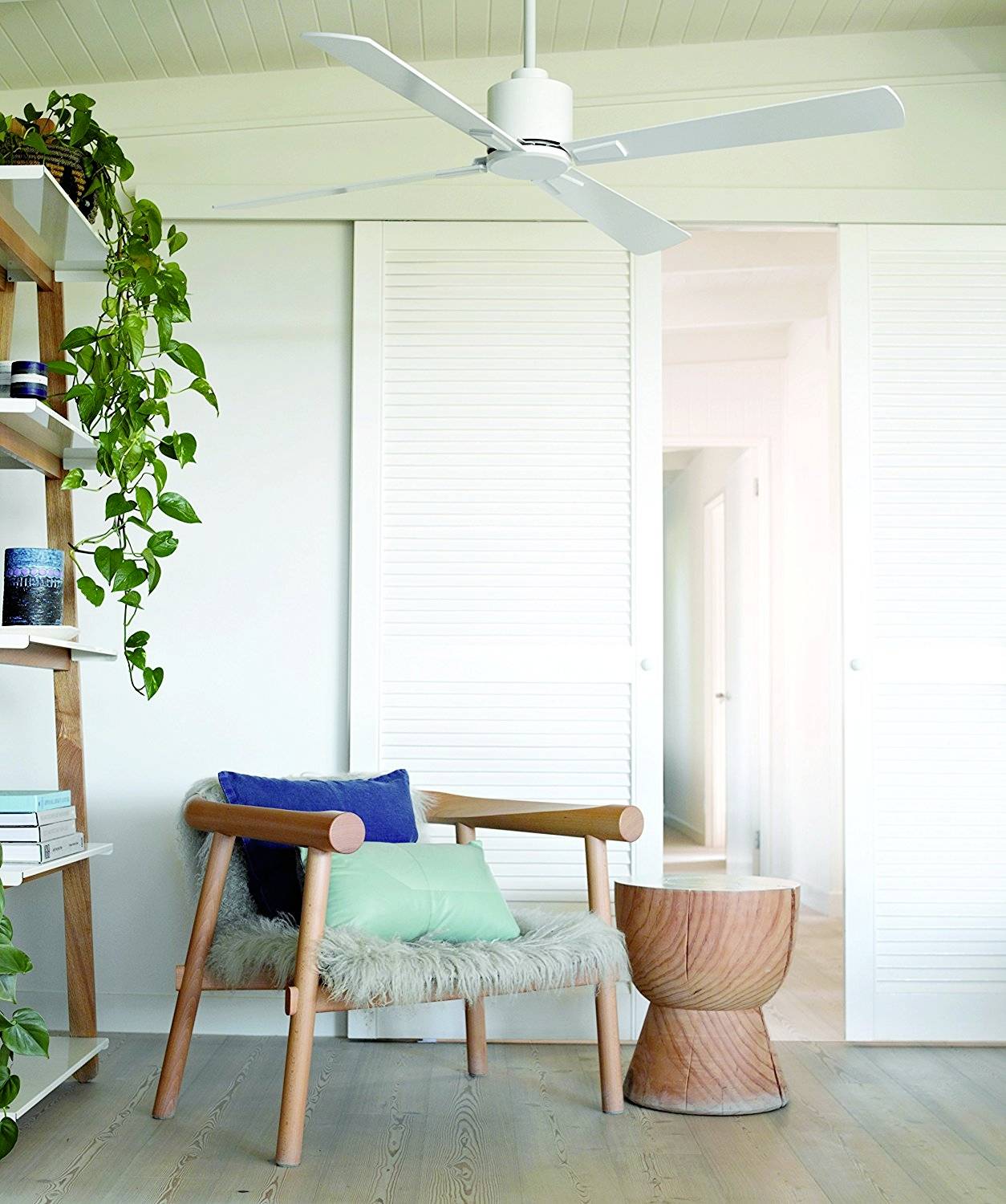 Helles Wohnzimmer mit einem Sessel, dekorativen Kissen, einem Holzhocker, einem Regal mit Pflanzen und einem Deckenventilator.