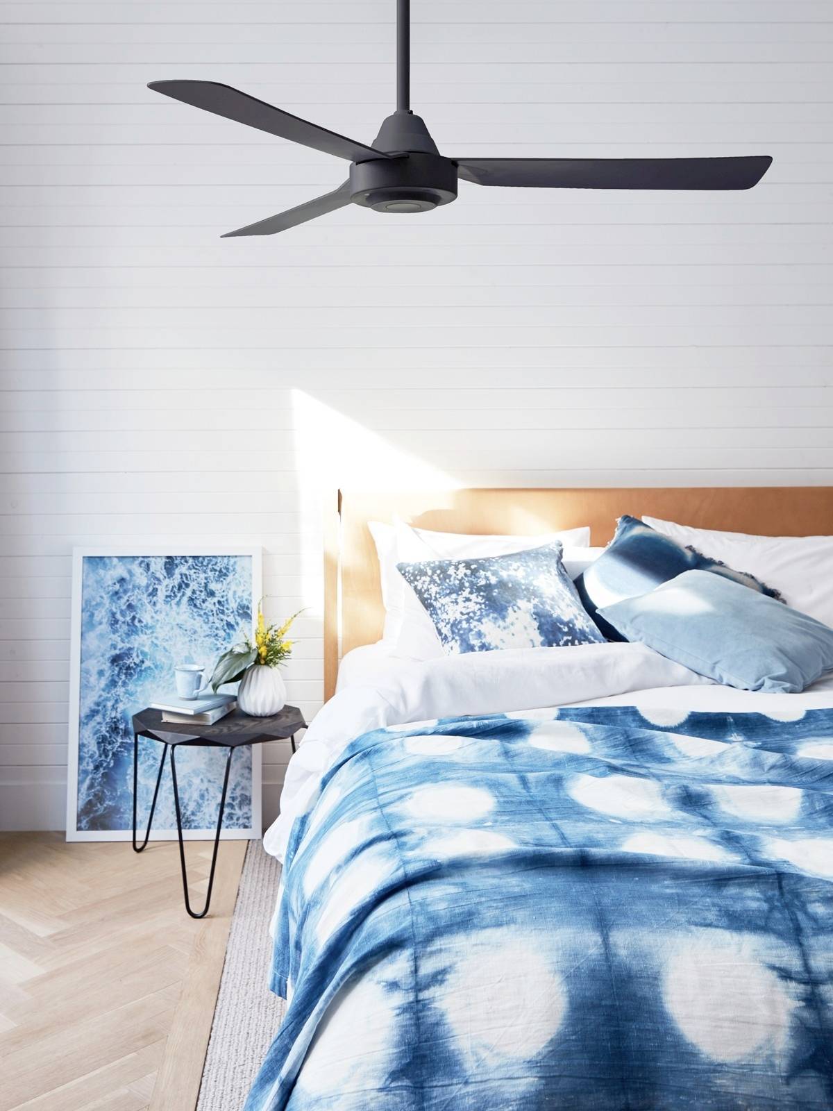 Ein modernes Schlafzimmer mit einem Bett, das blaue Batikbettwäsche und weiße Kissen hat. Ein kleiner Tisch daneben trägt eine Vase.