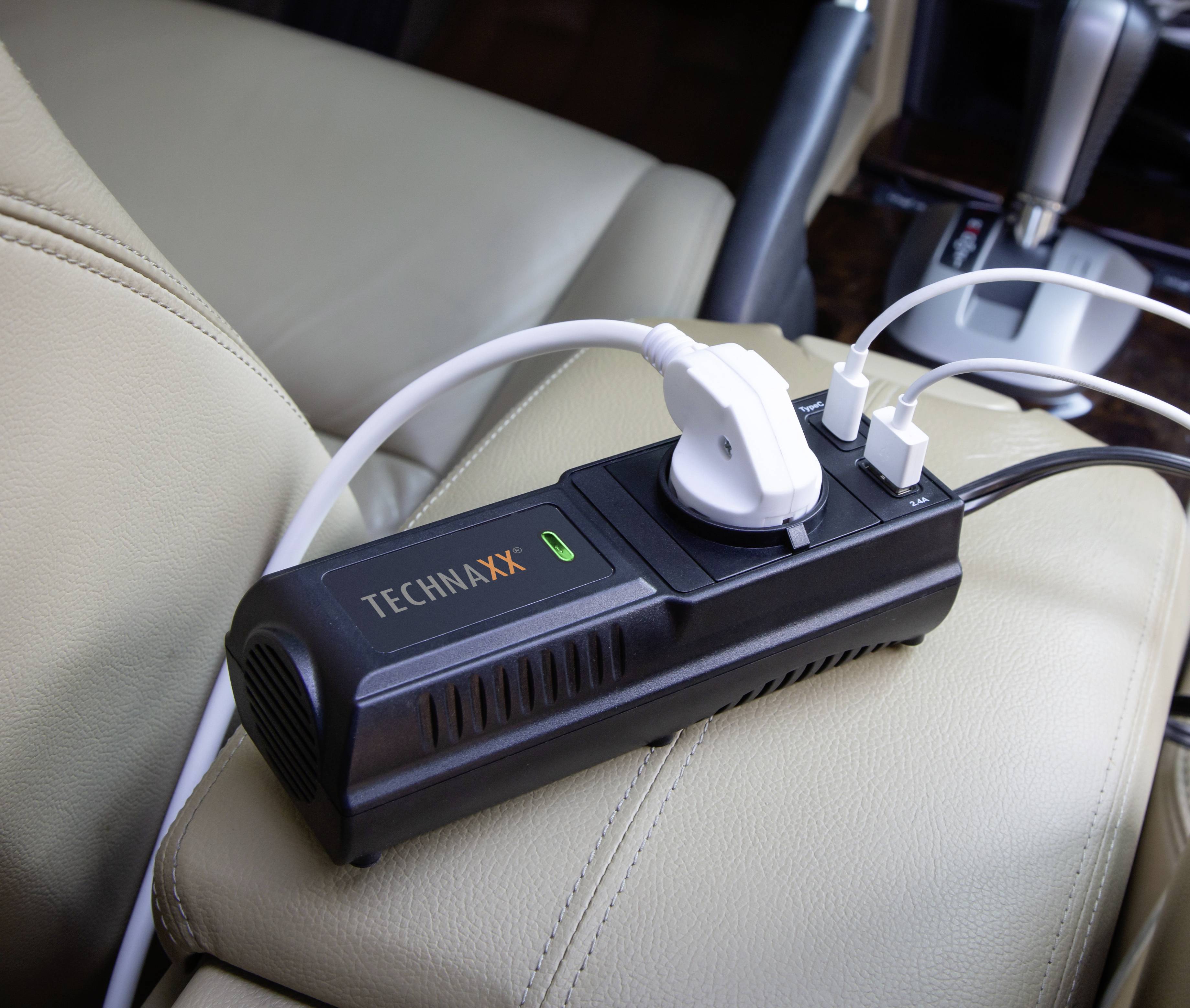 Stromadapter mit zwei USB-Anschlüssen und einer Steckdose, angeschlossen in einem Auto. Liegt auf einem beigen Ledersitz.