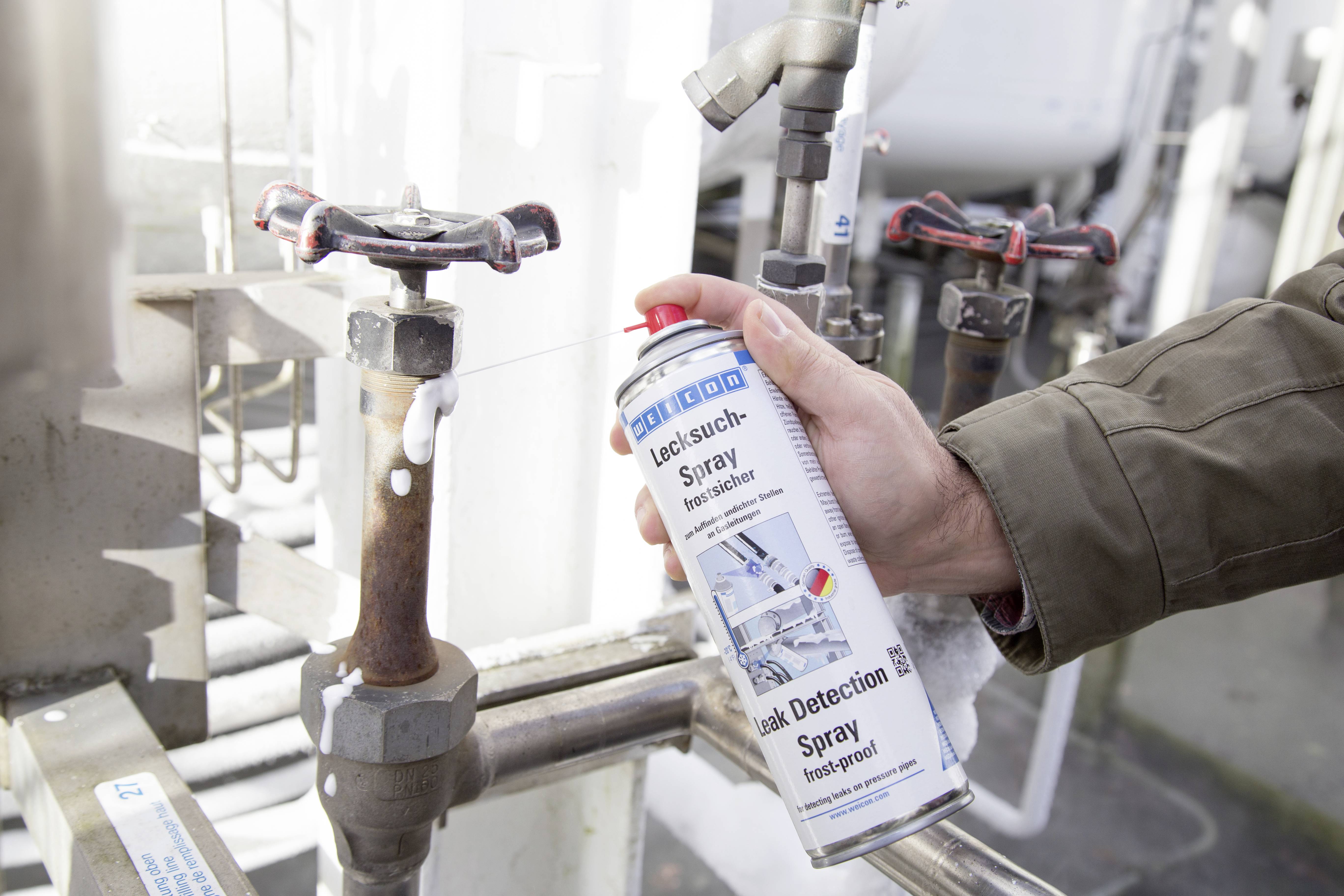 Person hält eine Dose Lecksuchspray und sprüht es auf ein Rohrventil in einer technischen Umgebung, um Lecks zu prüfen.