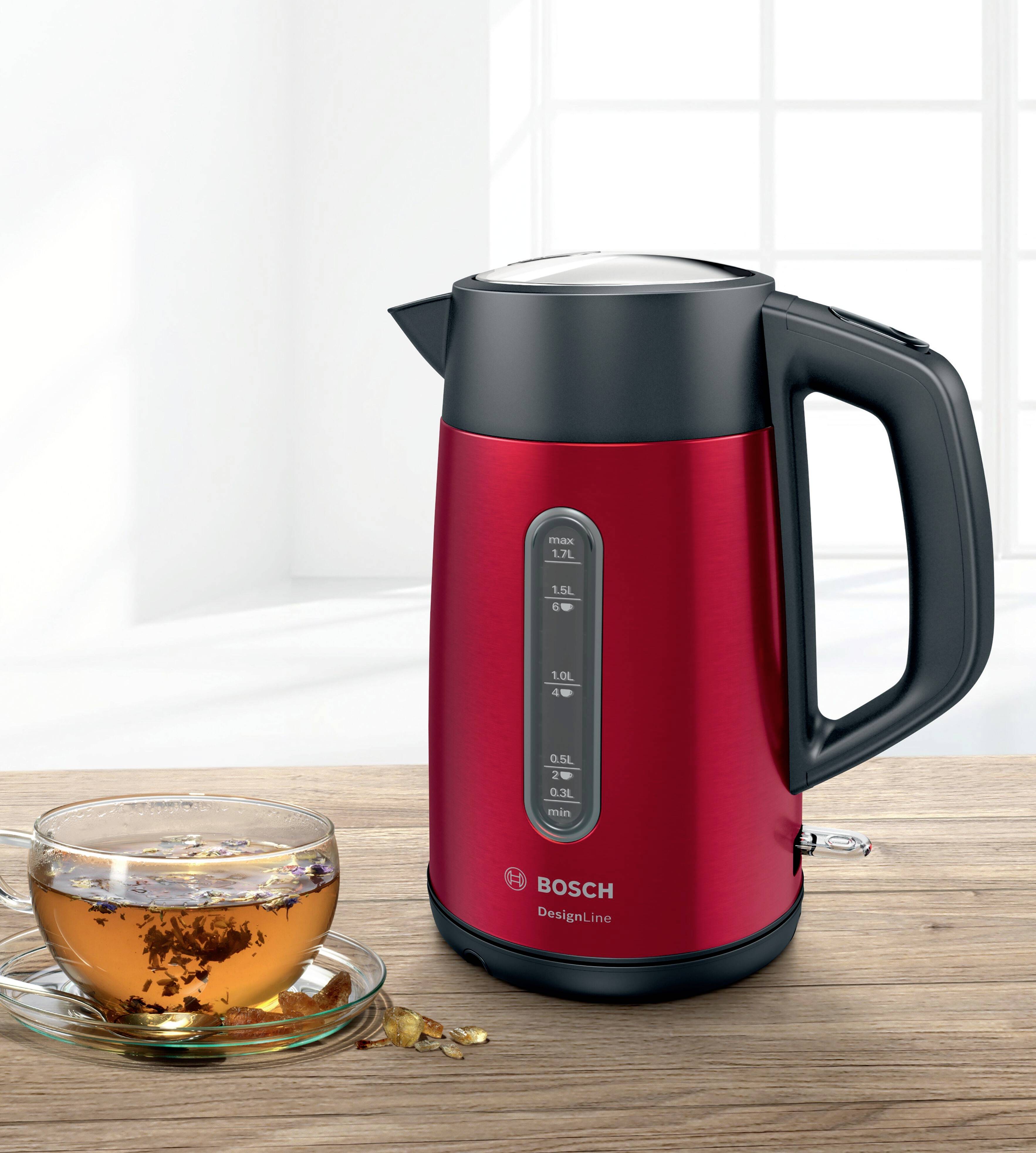 Ein roter Wasserkocher neben einer Tasse Tee in einem hellen Raum. Der Wasserkocher zeigt verschiedene Wasserstandsmarkierungen.