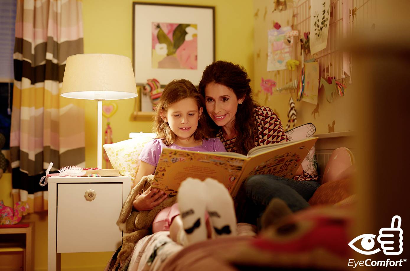 Eine Frau liest einem Kind in einem gemütlichen, beleuchteten Zimmer ein Buch vor. Beide schauen lächelnd auf die Buchseite.
